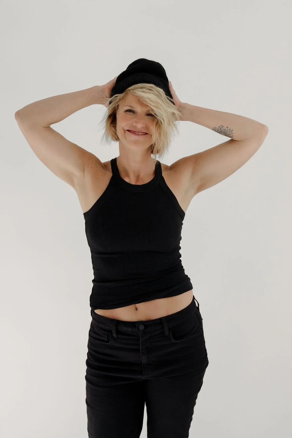 Smiling woman with short blonde hair, wearing a black tank top, black pants, and a black beanie, standing against a plain white background.