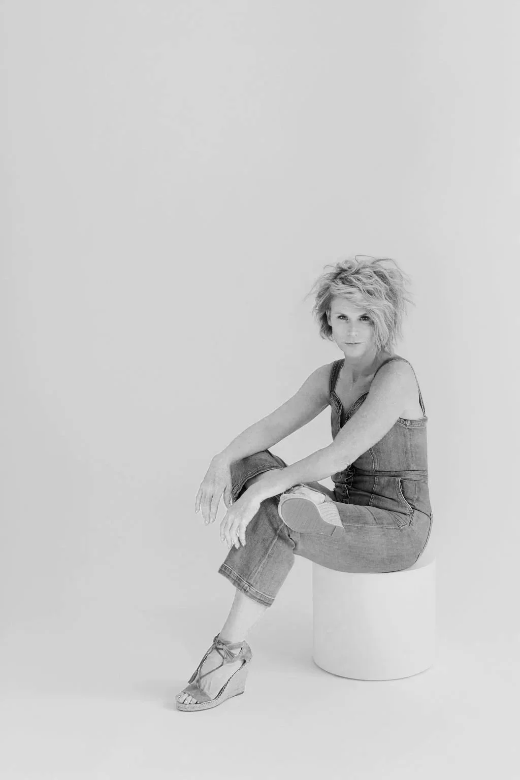 Black and white photo of a woman with tousled hair, sitting on a small round stool, wearing a sleeveless denim jumpsuit and high-heeled sandals, looking at the camera.