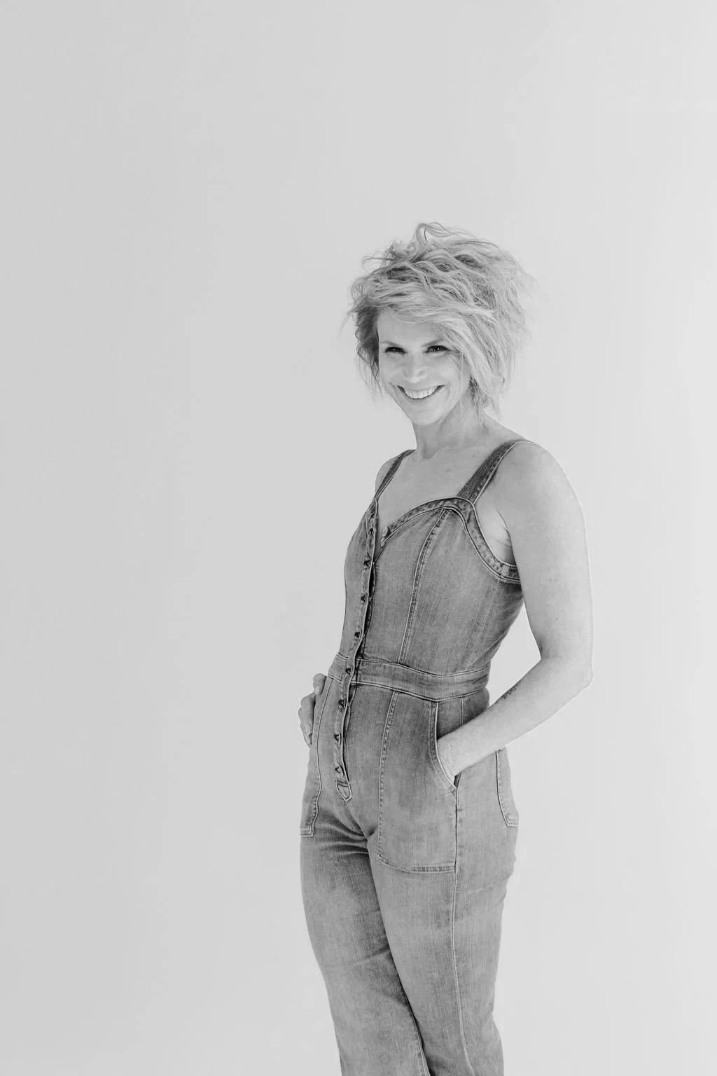 Black and white photo of a woman with short, wavy hair wearing a sleeveless denim jumpsuit with hands in pockets, smiling and standing against a plain background.