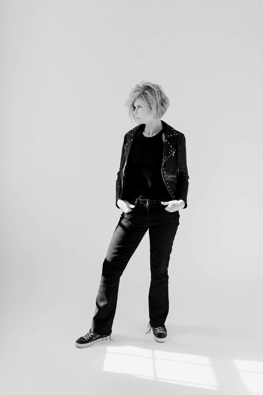 A woman with short, tousled hair stands against a plain light background. She is wearing a leather jacket with metal studs, a dark shirt, jeans, and sneakers, looking to her left with hands in her pockets.