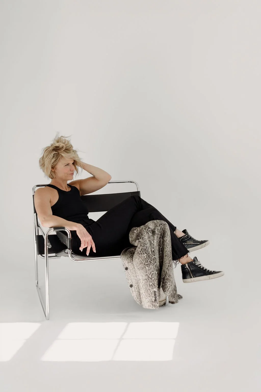 A woman with blonde hair in a black sleeveless top and black pants, sitting on a modern chair against a plain white background, with a snake-print jacket draped over her legs.
