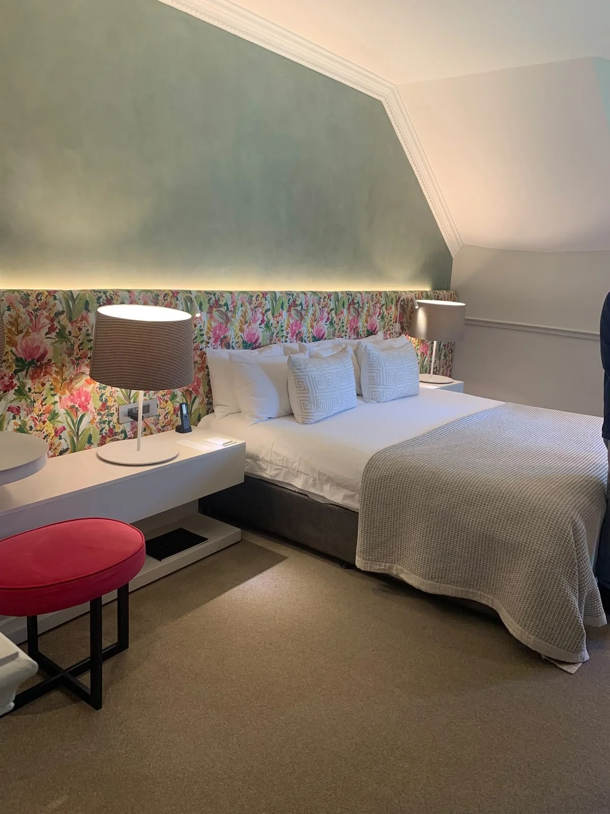 A hotel room with a bed that has a floral upholstered headboard, white sheets, and decorative pillows. There are two lamps on white nightstands on either side of the bed. A red stool and a white desk are nearby. The room features a sloped ceiling with crown molding and soft lighting.