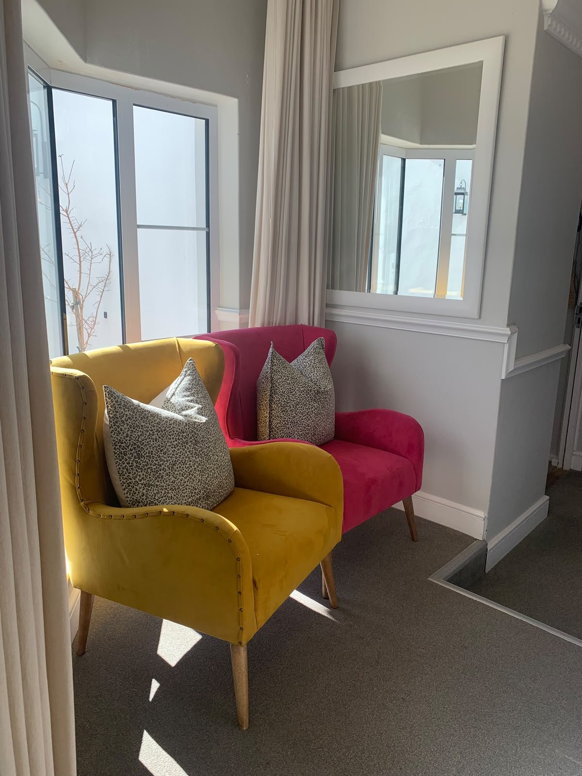 A corner of a room with two velvet armchairs, one yellow and one pink, each with leopard print pillows. A large mirror is mounted on the wall next to beige curtains covering a window, allowing natural sunlight to illuminate the space.