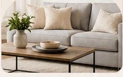 Living room with a light gray sofa, beige pillows, a wooden coffee table, a white vase with green plants, and a neutral rug.