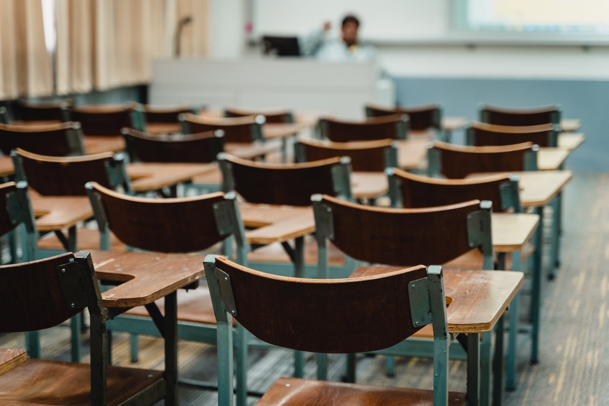 Empty classroom with multiple wooden desks and chairs arranged in rows, with a teacher standing near a whiteboard at the front of the room.