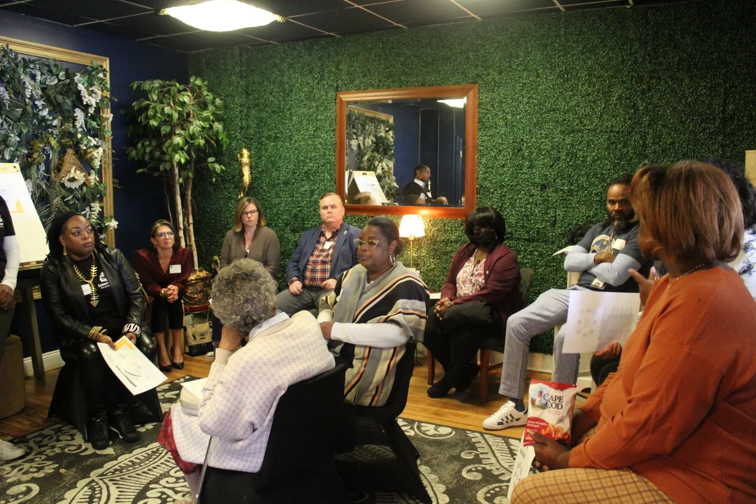 Group of diverse people sitting and listening to a speaker in a room with green leafy wall, mirror, and decorative plants.