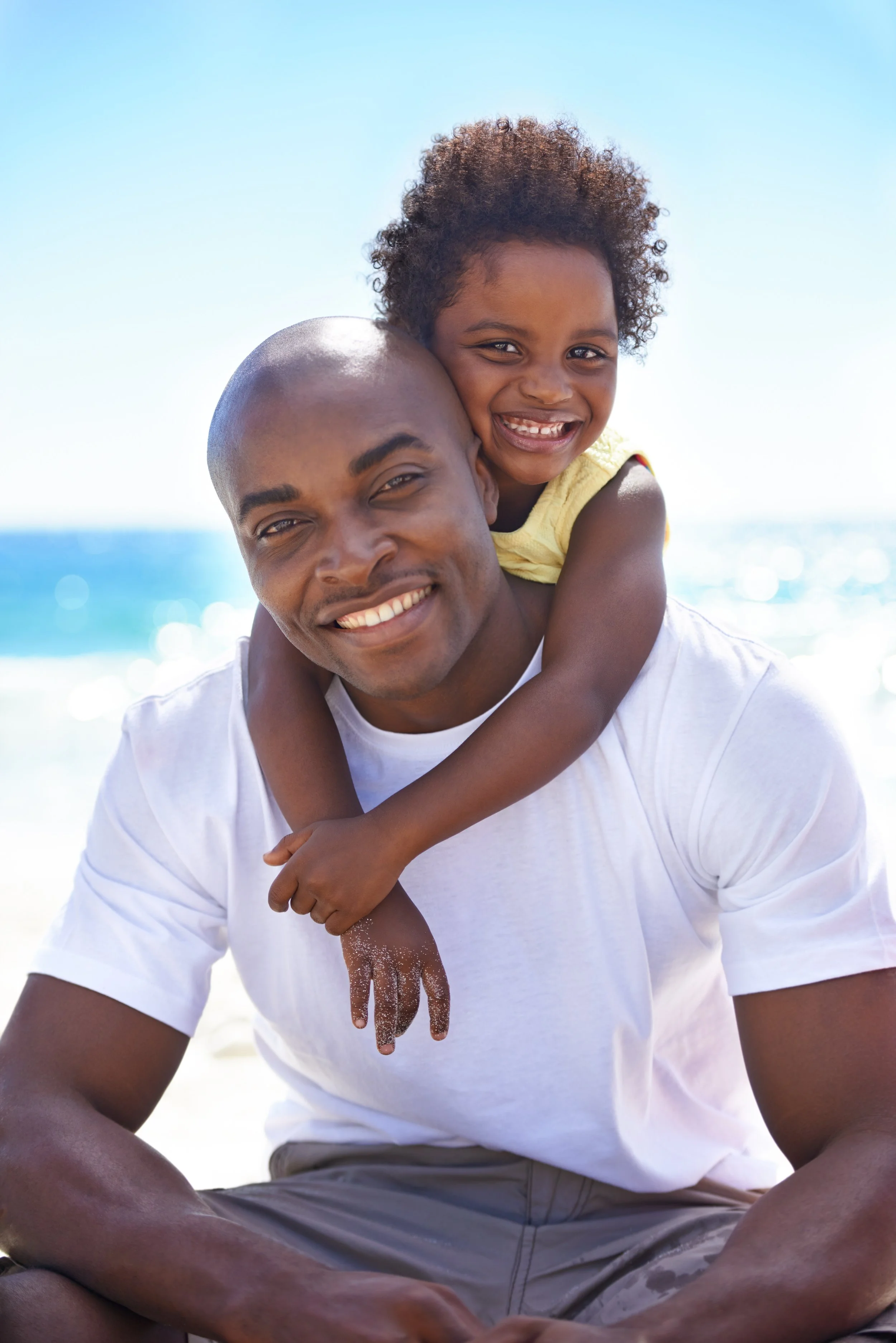 A man with a shaved head and a white t-shirt sitting on the beach, smiling, with a young girl with curly hair on his back, also smiling, and they are near the ocean under a clear blue sky.