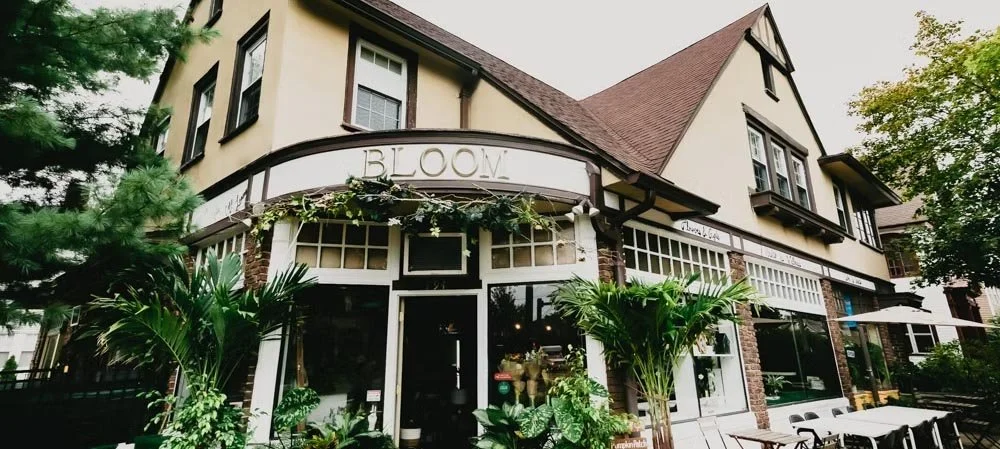 A two-story building with a business named 'Bloom,' featuring large glass windows and outdoor seating, surrounded by greenery and trees.