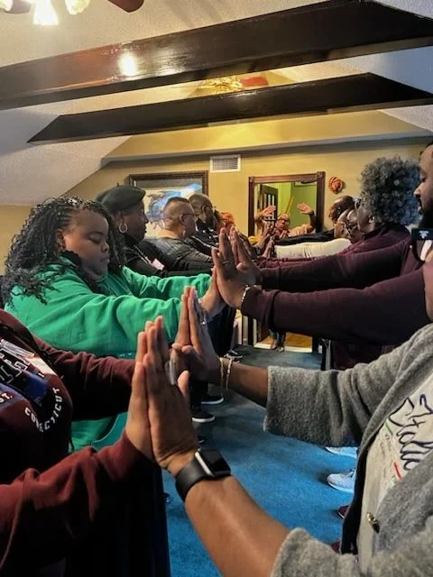 Group of diverse people practicing a synchronized prayer or meditation with hands pressed together, standing shoulder to shoulder in a room.