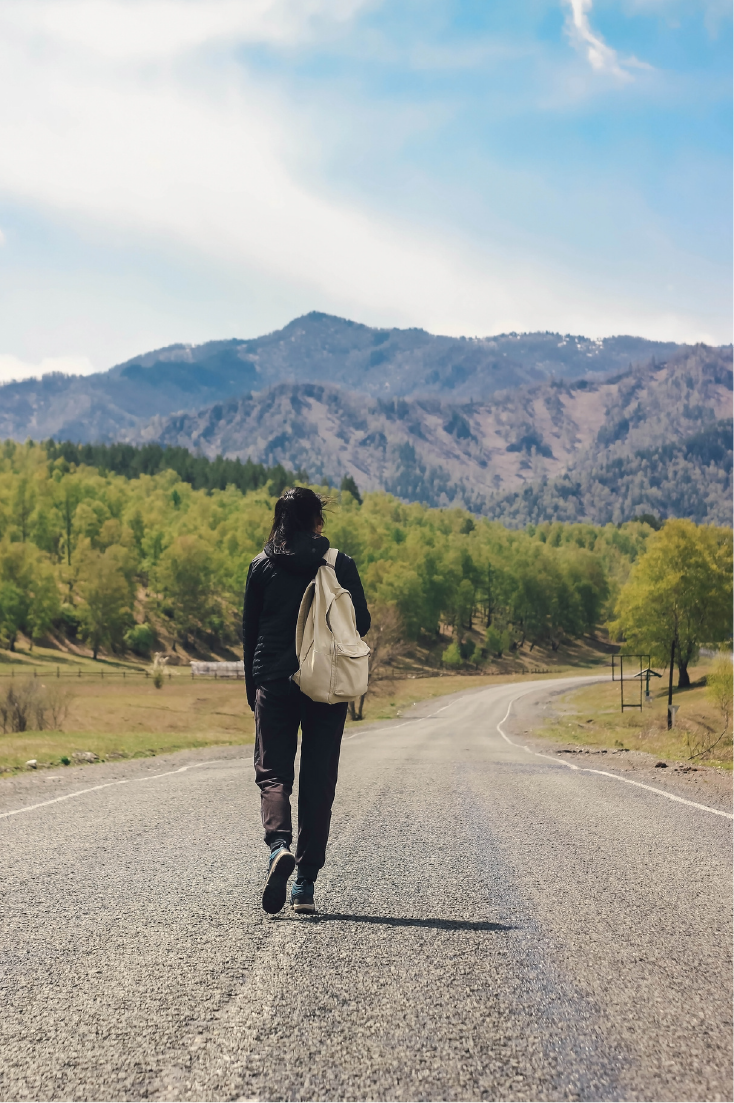 A person walking on a rural road with mountains and trees in the background on a sunny day.
