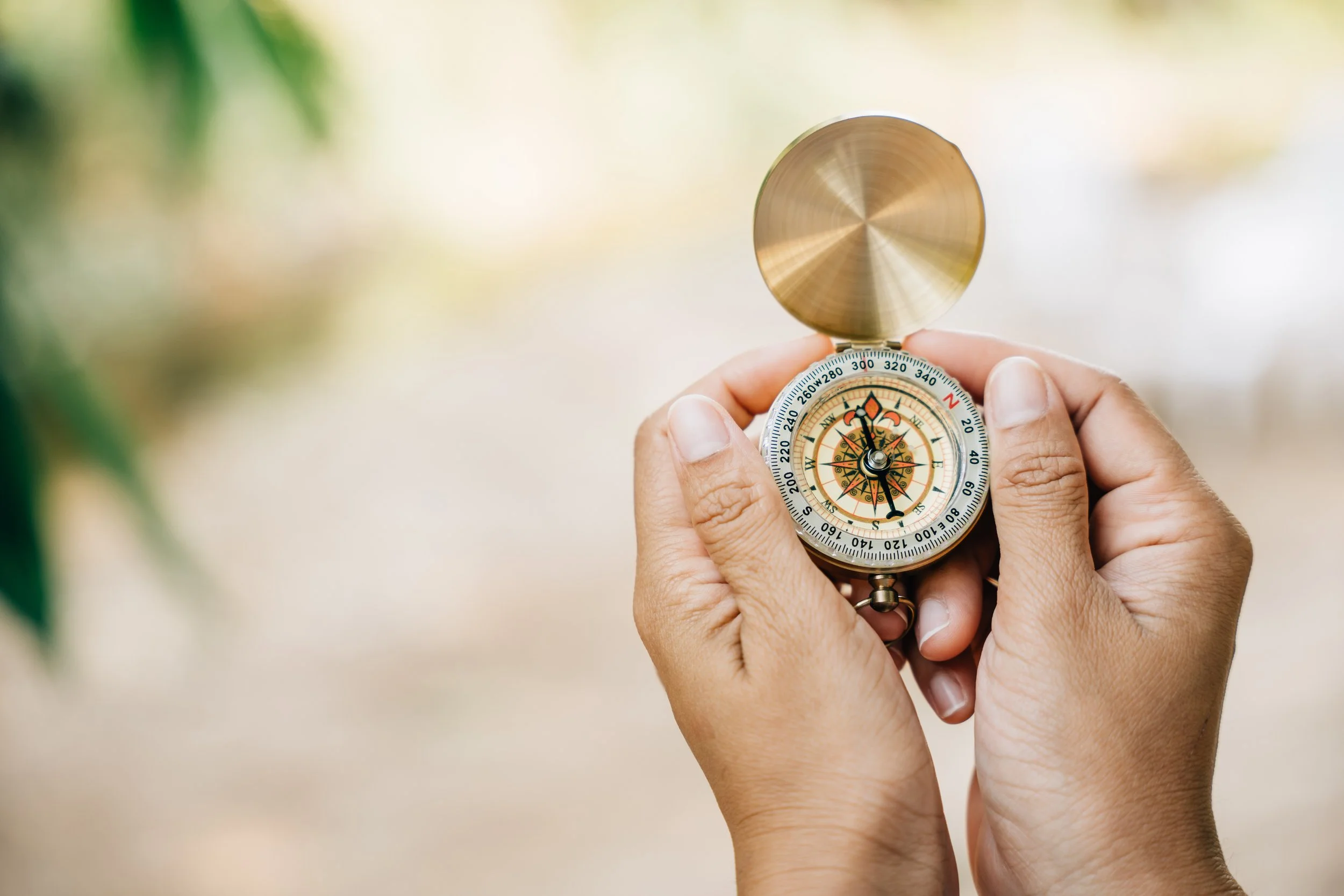 A person holding a vintage compass with a metal lid, pointing outdoors with a blurred background of green foliage and light-colored ground.