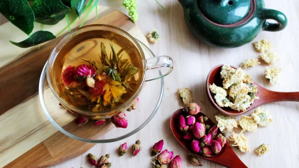 A glass cup of herbal tea with dried flower petals inside, placed on a clear glass saucer on a wooden surface. Surrounding it are two wooden spoons filled with dried flowers or buds, a green teapot, and some green leaves in the background.