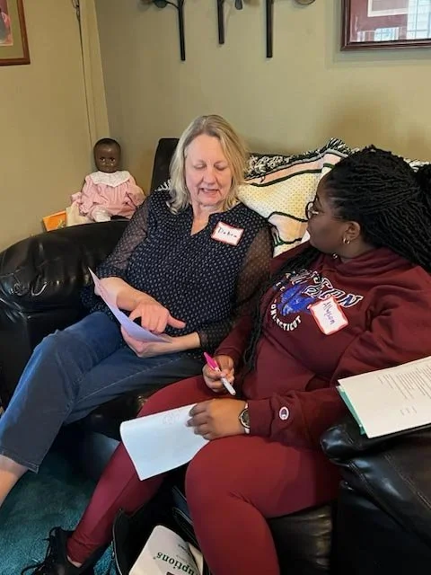 Two women sitting on a black leather couch, engaged in conversation, with a doll sitting in the background.