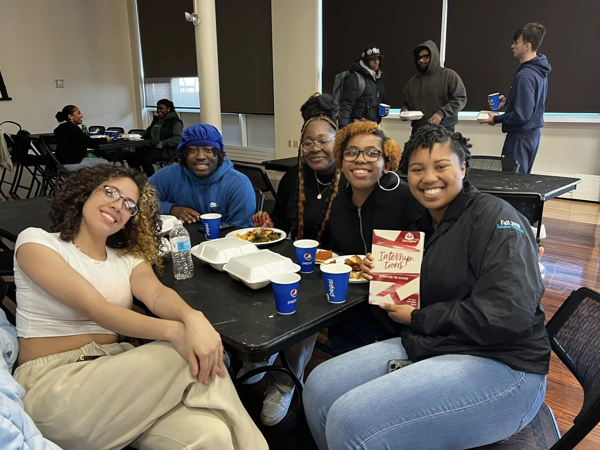 Group of diverse young adults sitting at tables with food and drinks, smiling and enjoying each other's company in a social setting, with some standing and talking in the background.