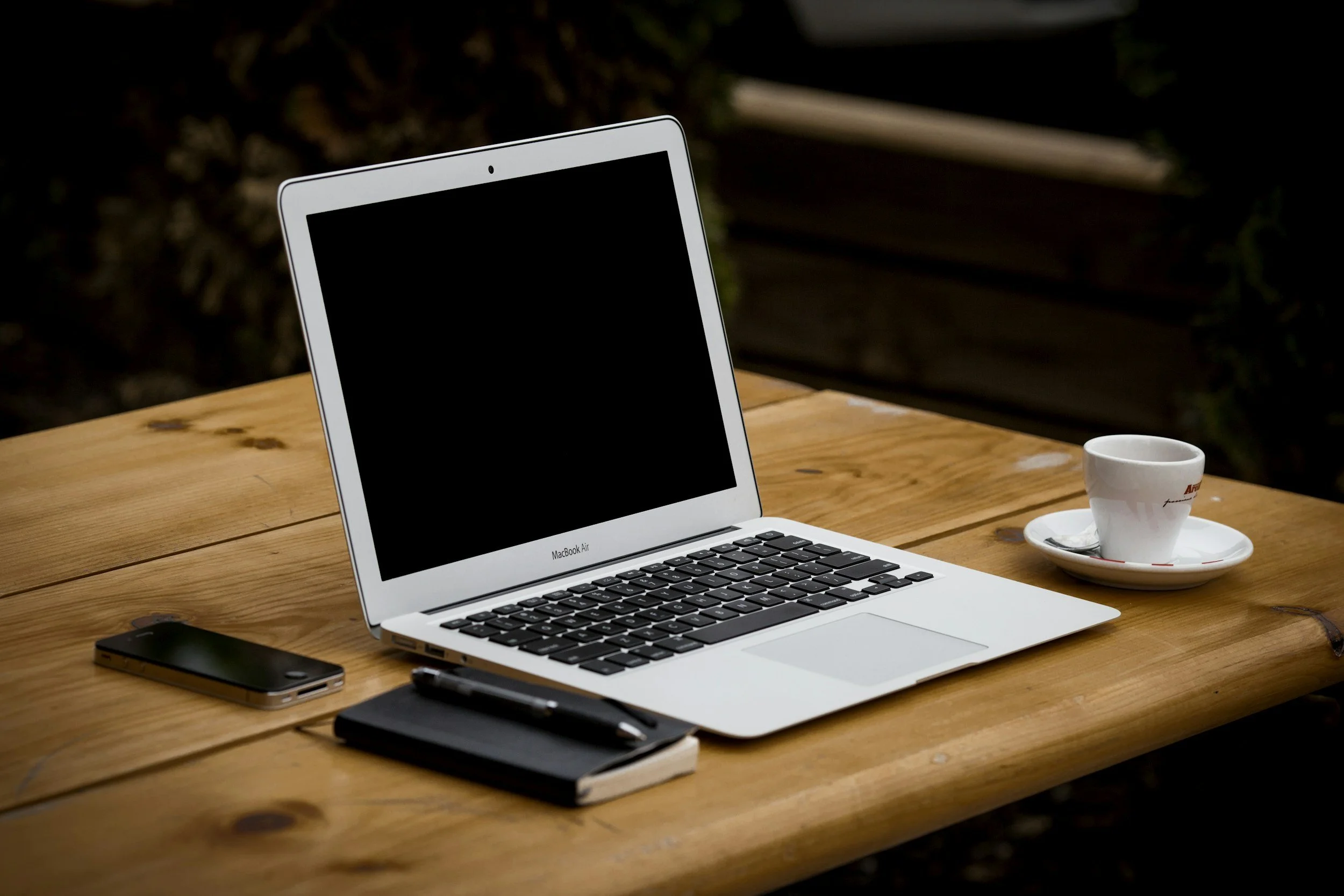 A MacBook Air laptop on a wooden table, with a coffee cup on a saucer, a black notebook, a pen, a smartphone, and a black object nearby, in an outdoor setting.