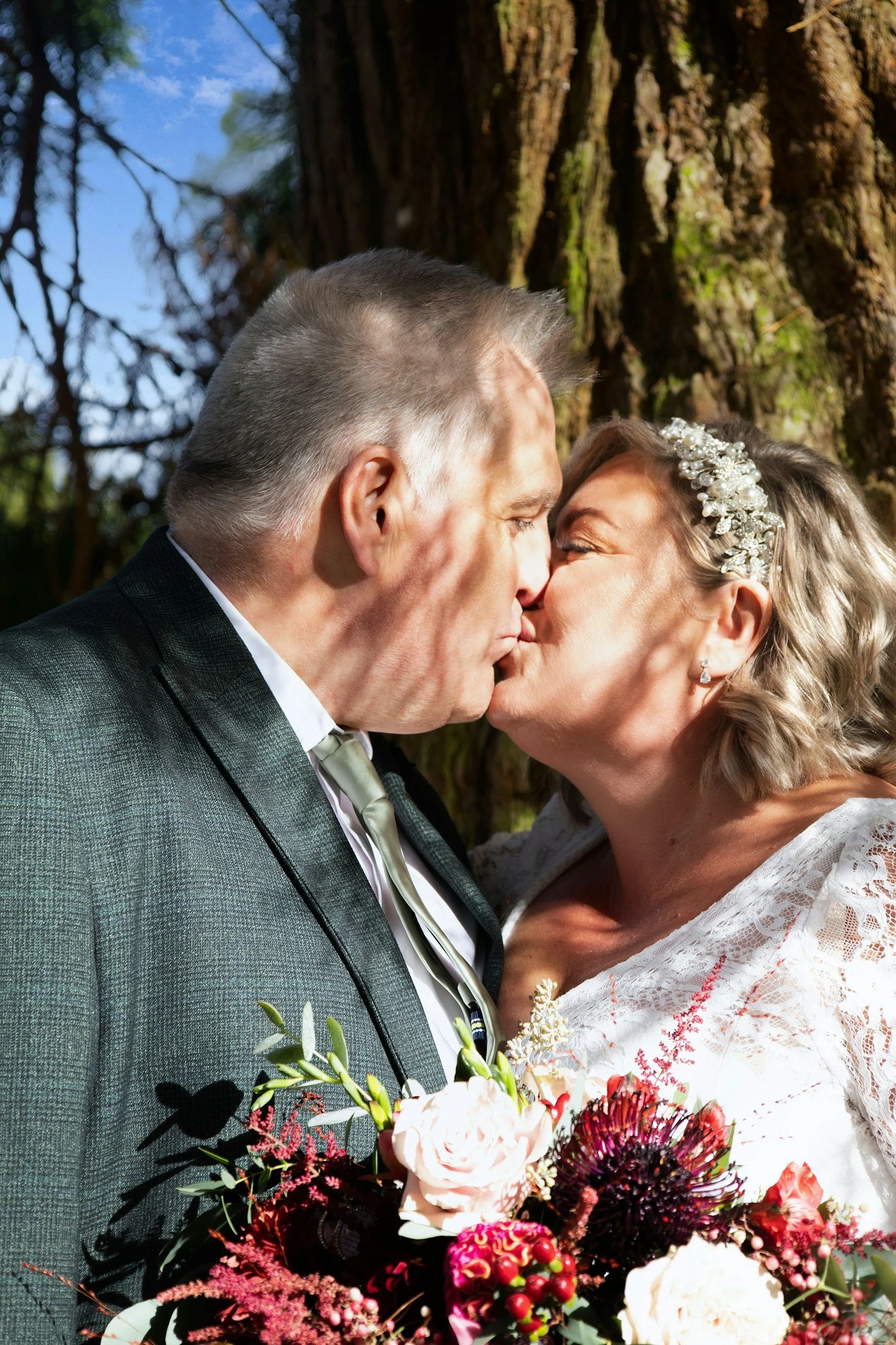 A kissing couple at a wedding, outdoors near a large tree, with the woman holding a bouquet of flowers.