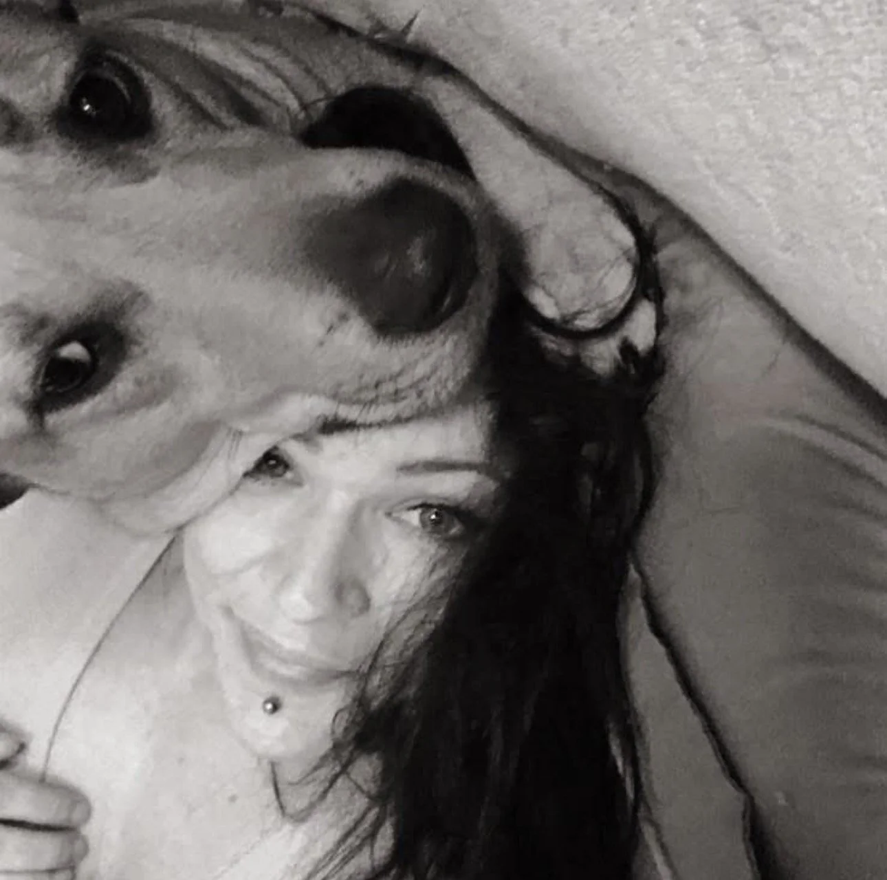 A person with long dark hair lying next to a large dog with light fur, both resting on a surface. The person's face is close to the dog's face, and they are looking at the camera.