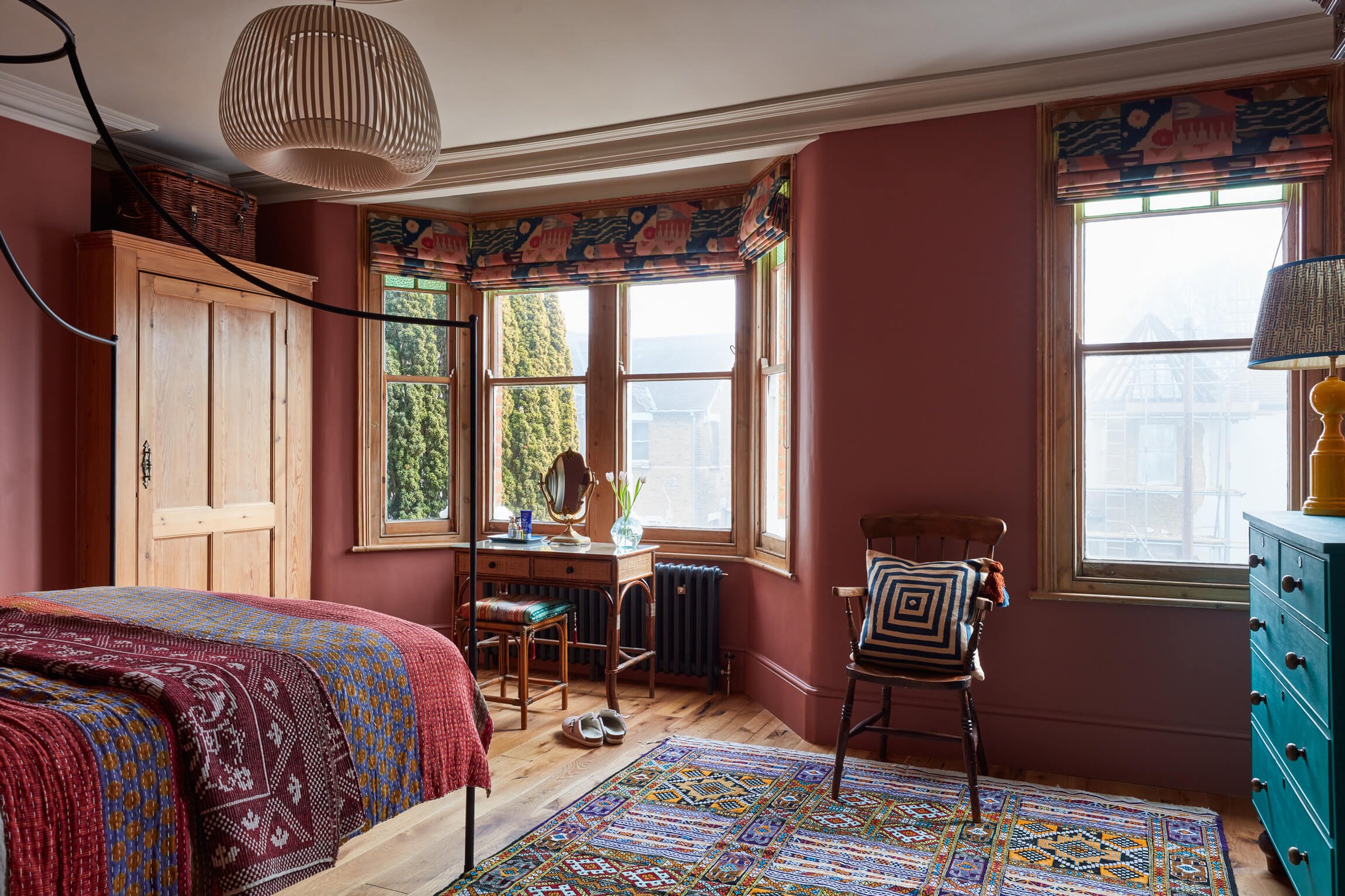 Interior view of a cozy bedroom with large bay windows, wooden furniture, colorful patterned bedding, a vintage dresser, a zebra print cushion, and a vibrant area rug.