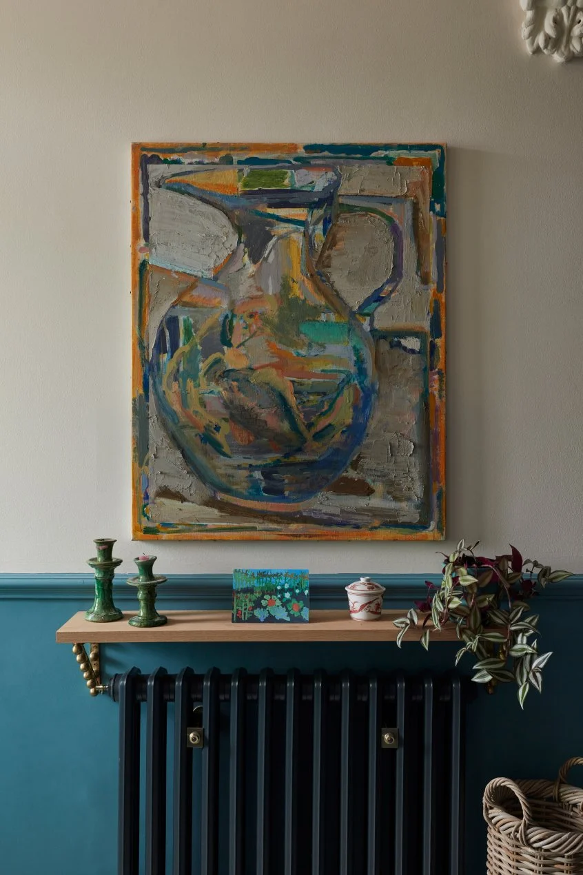 A colorful abstract painting of a pitcher hangs on a plain wall above a wooden shelf decorated with green candlesticks, a small floral painting, a white jar with a red ribbon, and a plant with green and purple leaves. The shelf is mounted above a dark radiator and a wicker basket is visible at the bottom right.