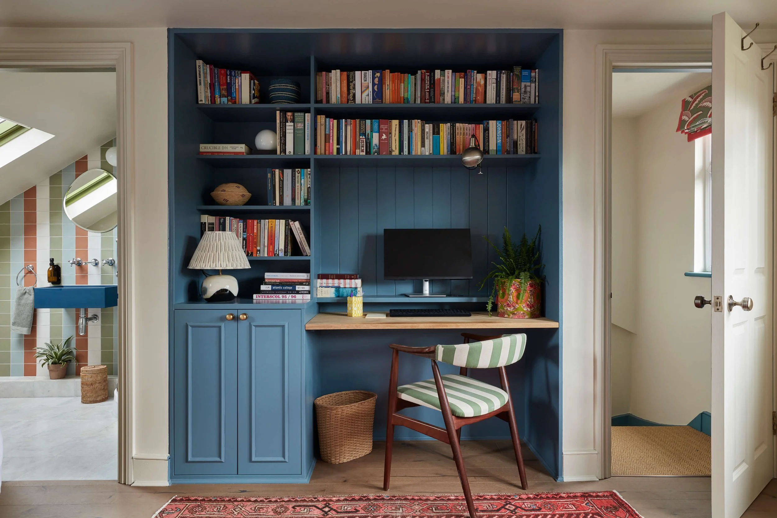 Blue built-in bookcase and desk nook with a striped chair and table lamp, with doorways leading to a colourful tiled bathroom and hallway.