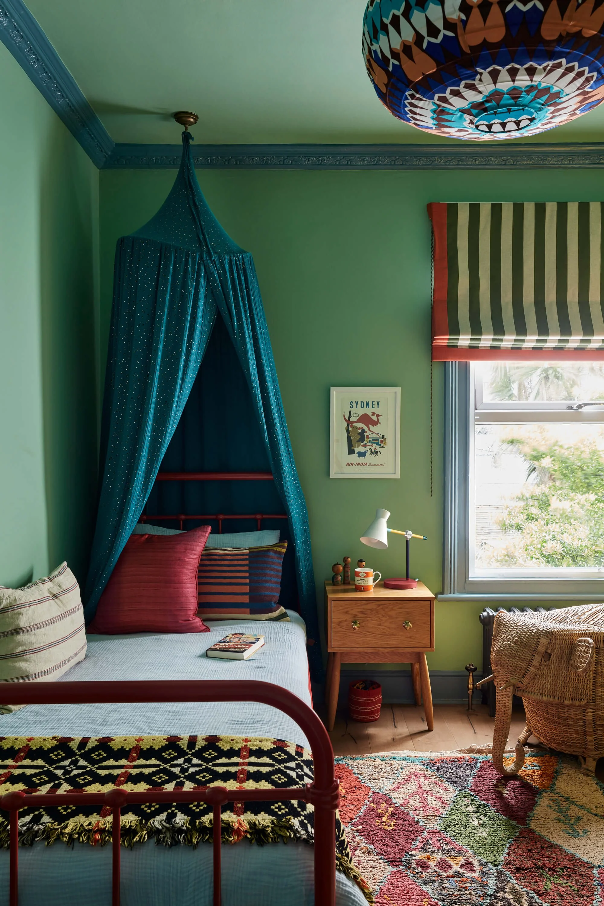 A bedroom with green walls, decorated with colorful textiles and furniture. A bed with a red metal frame is in the foreground, with striped and solid pillows on a white bedspread. A small wooden nightstand holds a pink desk lamp, a mug, and small decor items. To the right, there is a woven chair with a cushion, a window with green and white striped Roman shades, and a multicolored patterned rug on the wooden floor. A framed print labeled 'Sydney' hangs between the window and the bed, and a blue canopy drapes from the ceiling.