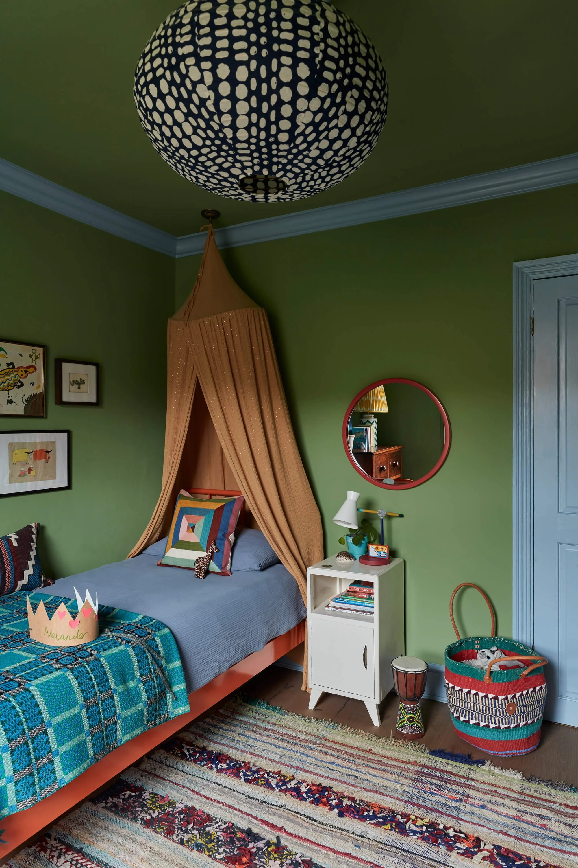 A bedroom with green walls, a bed with a blue and green plaid bedspread, and a canopy over the bed. There are framed pictures on the wall, a red-framed mirror, a small white nightstand with books and decor, and a colorful woven basket and drum on the floor. A striped area rug is partially visible.