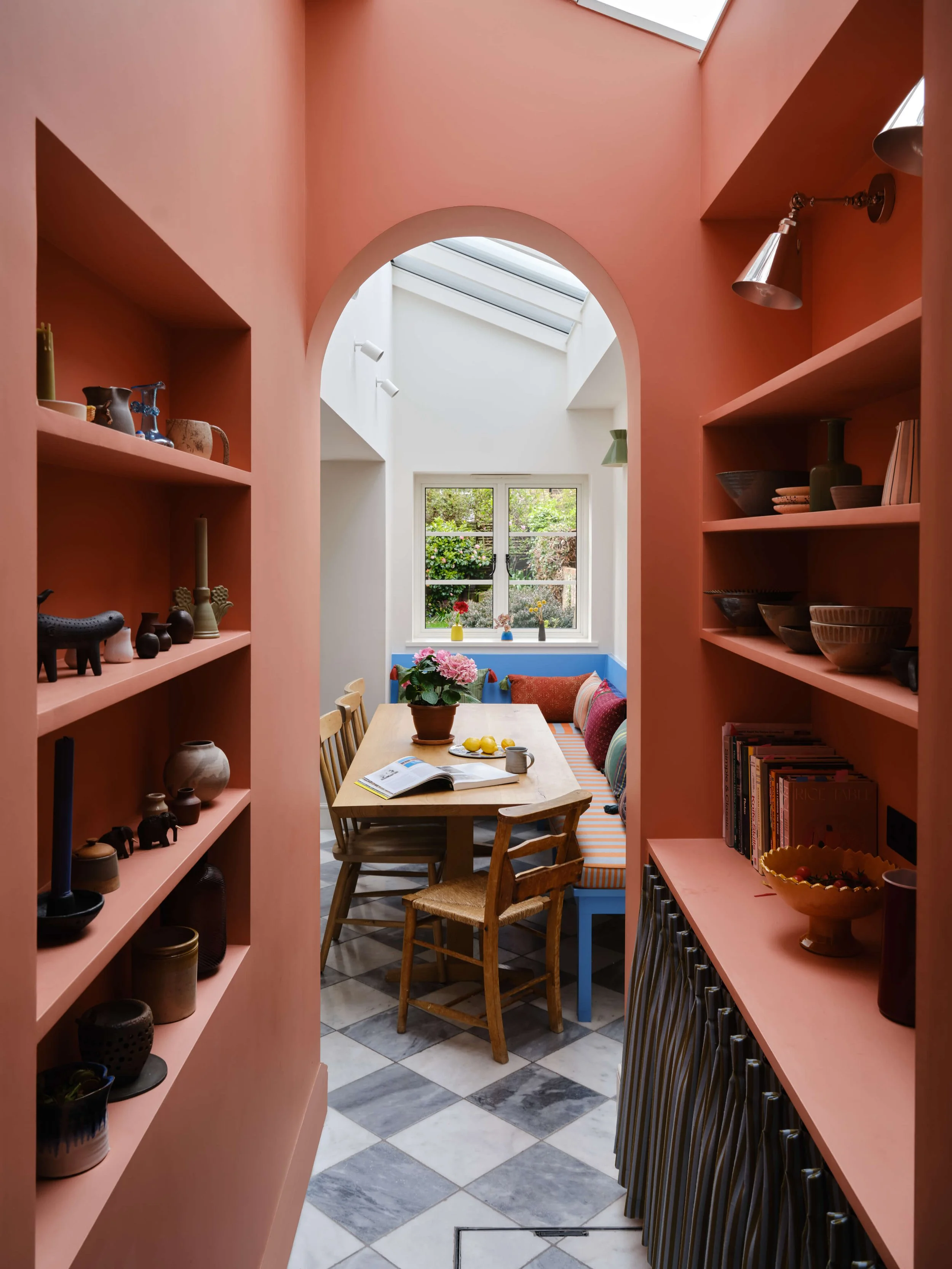 A cozy dining nook with pink built-in shelves filled with decorative pottery and books, a wooden dining table with a open magazine, and a window with a garden view, illuminated by skylight and wall-mounted lights.