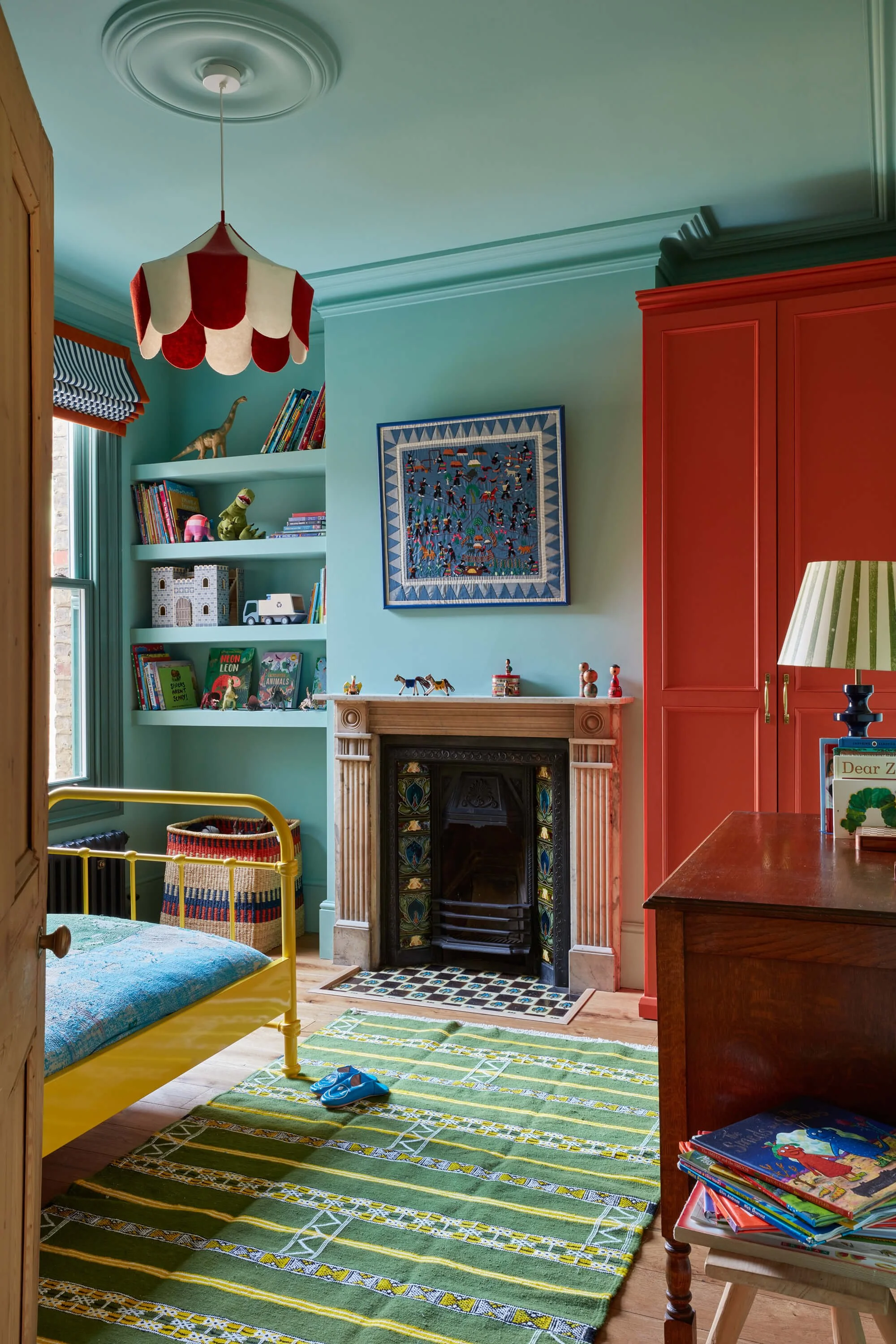 A vibrant children's bedroom featuring a yellow bed, a colorful striped area rug, a fireplace, built-in bookshelves filled with books and toys, a red wardrobe, and a framed artwork on the wall. The room has a ceiling light with a red and white scalloped lampshade, and a window with a striped window shade.