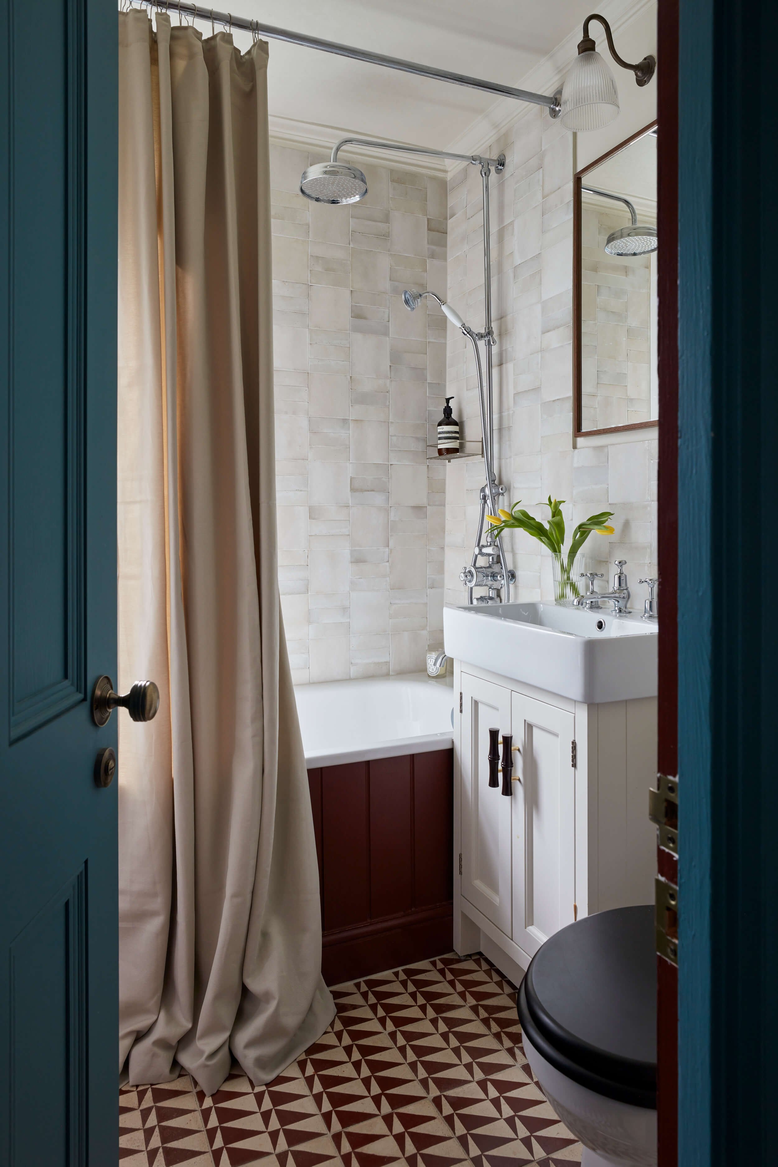 Bathroom with shower area, beige tiles on the wall, a brown and white patterned tile floor, a white vanity with a small mirror, a vase with yellow lilies, a wall light, and a beige shower curtain.