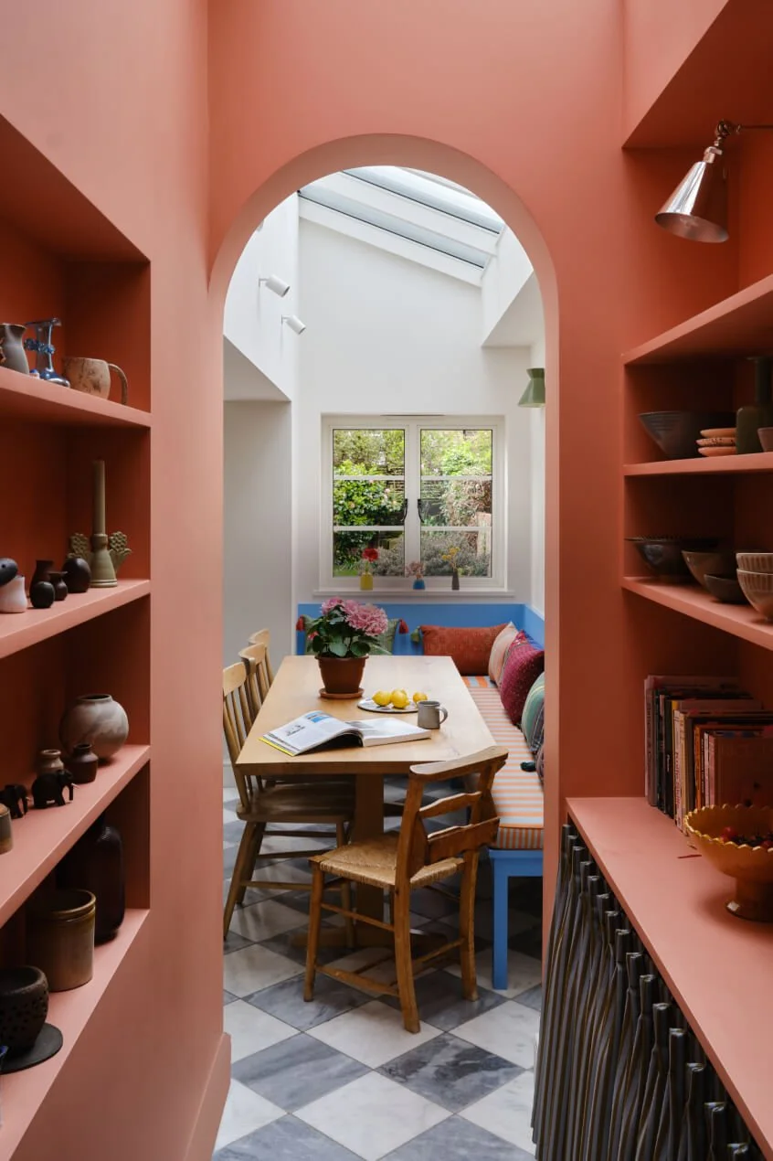 A cozy dining nook with a wooden table, surrounded by chairs and a built-in colorful bench. The space is lit by a large window and skylight, with pink walls and open shelving displaying pottery and books.