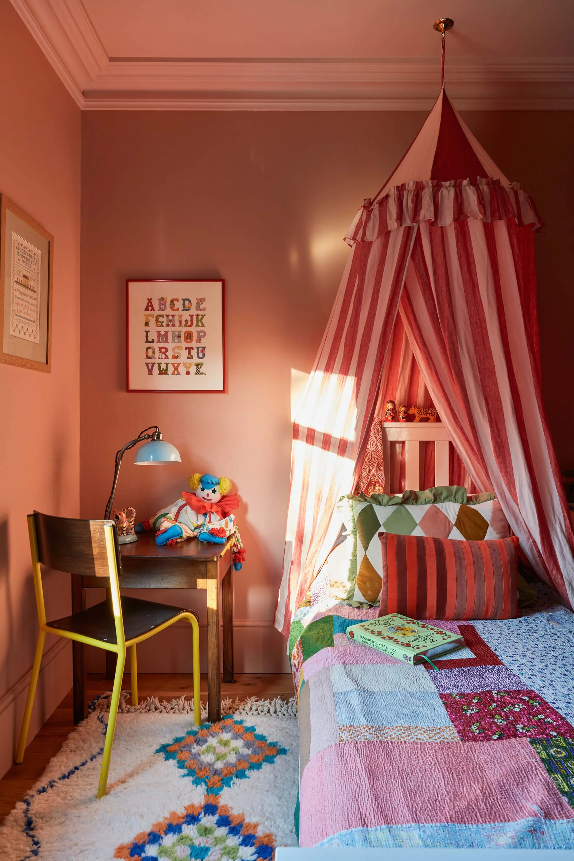 A cozy children's bedroom with a canopy bed featuring red and white striped curtains and a colorful patchwork quilt. There is a wooden desk with a yellow chair, a classic green banker lamp, toys, and a book on the bed. The walls are decorated with framed alphabet and sewing-themed artwork. A plush flower-shaped rug is on the wooden floor.