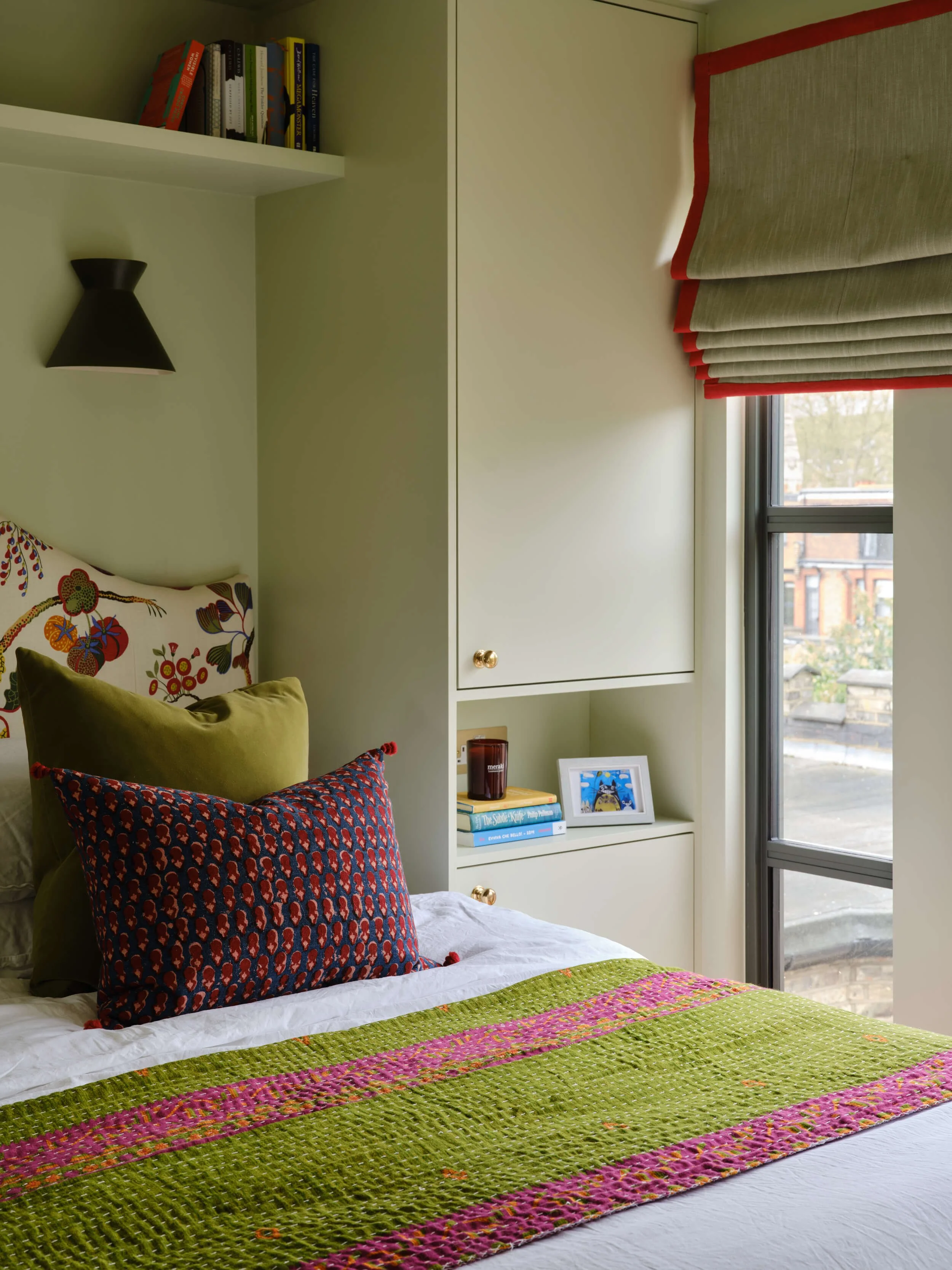 Bedroom with a colourful patterned bedspread and cushions, built-in shelf and a window with a striped blind.