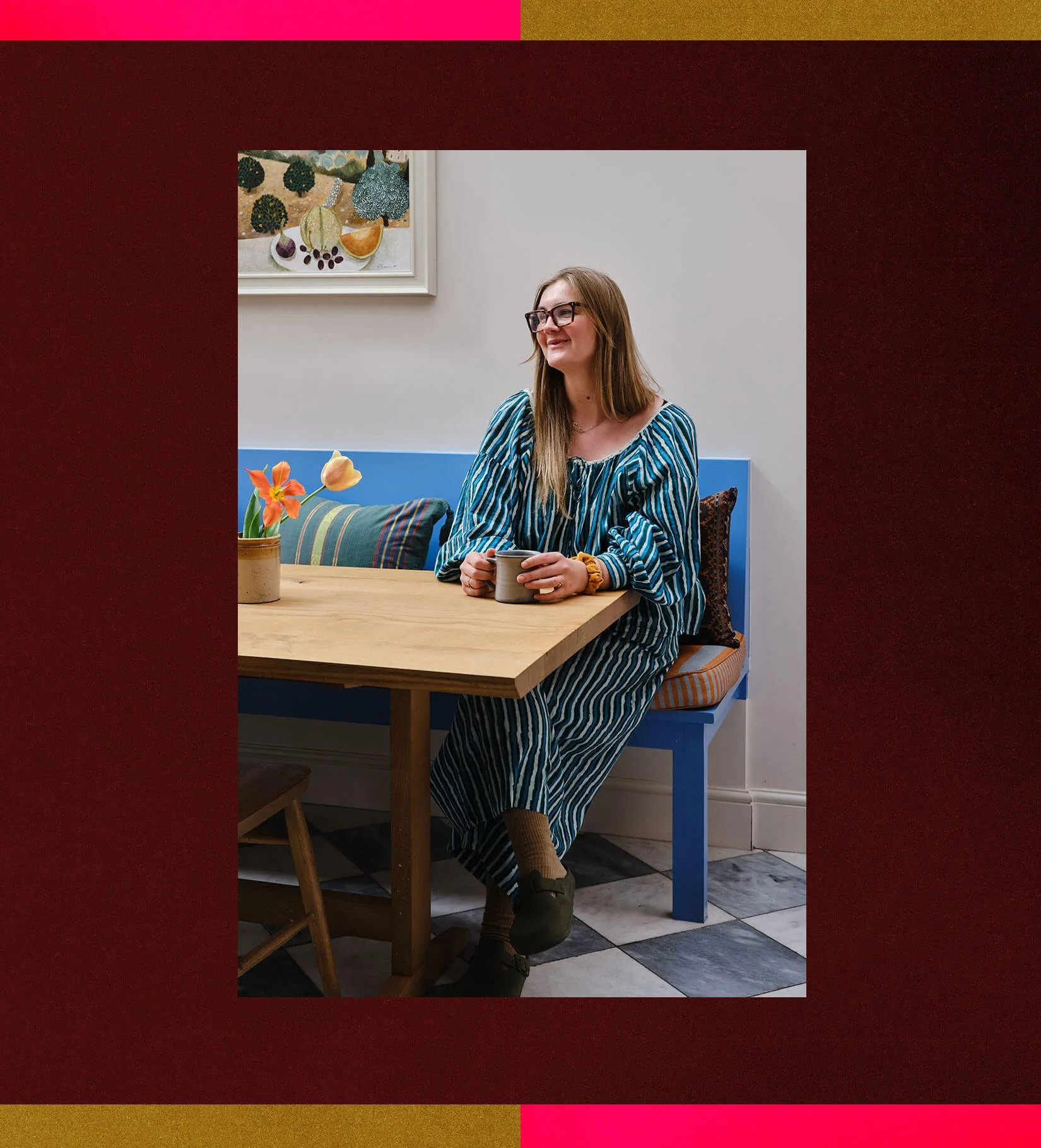 Sophie Gunning, founder of Project Home, sitting at a wooden table on a blue bench with tulips and framed artwork behind her.