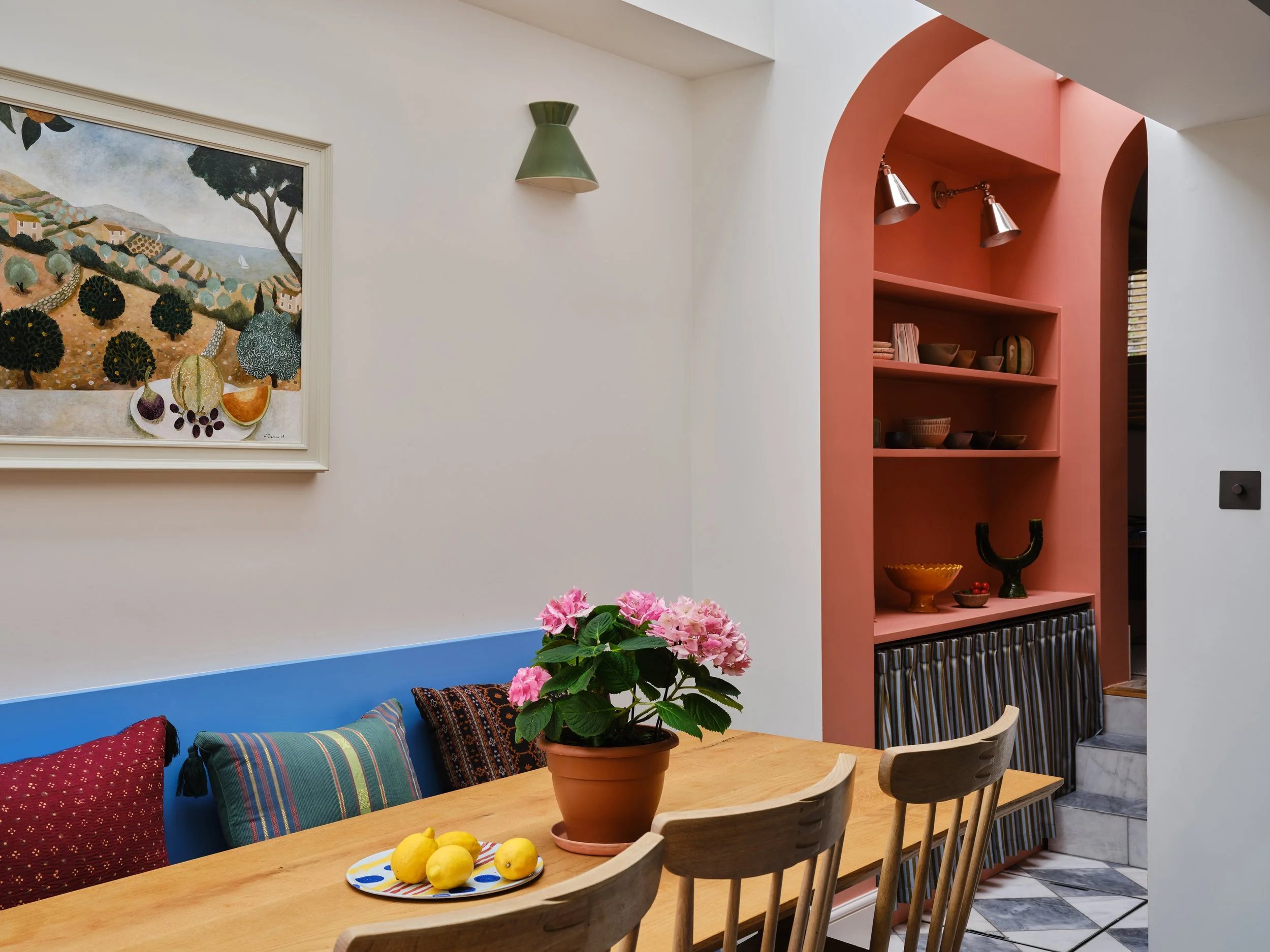 Interior of a dining area with a wooden table, colorful pillows on a bench, a pot with pink flowers, a plate of lemons, pink and white walls, a framed landscape painting, and a built-in pink shelving unit with dishes and decor.
