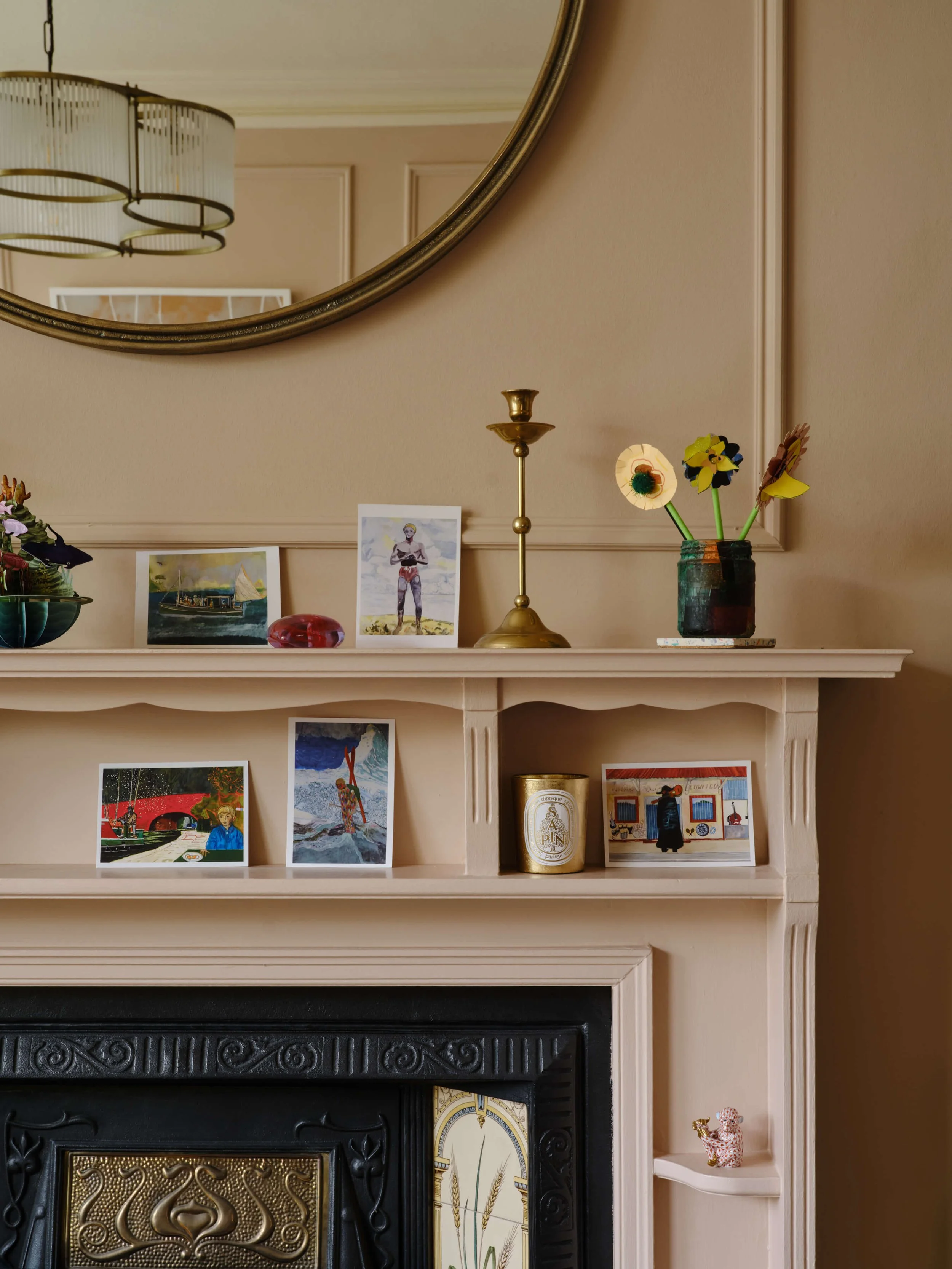 White fireplace mantel styled with framed artwork, a gold candle holder and flowers beneath a large round mirror.