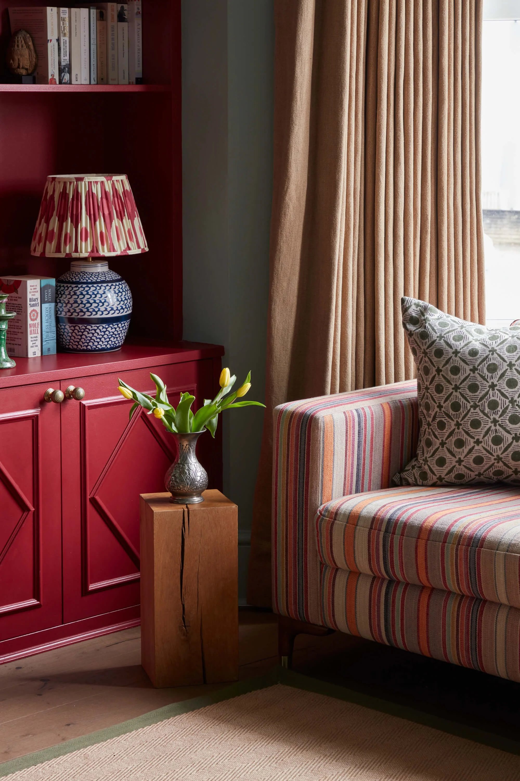 A cozy living room corner with a red cabinet, a blue and white patterned table lamp, books on the red shelf, a small wooden side table with a silver vase holding yellow tulips, a striped upholstered chair with a patterned pillow, beige curtains, and a window letting in natural light.
