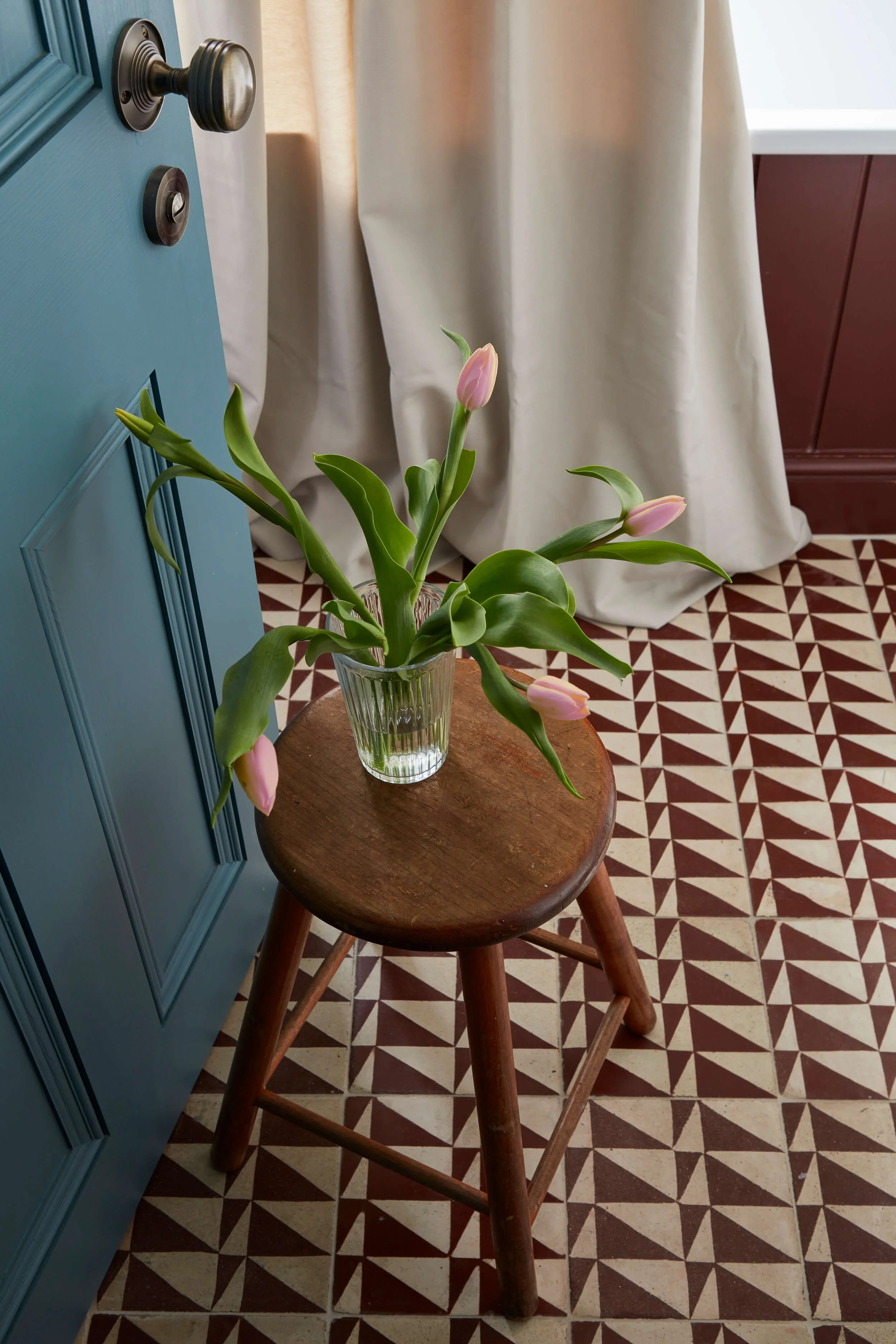 A wooden stool with a glass vase containing pink tulips on top. The background features a patterned floor, a blue door, a beige curtain, and a red wall.
