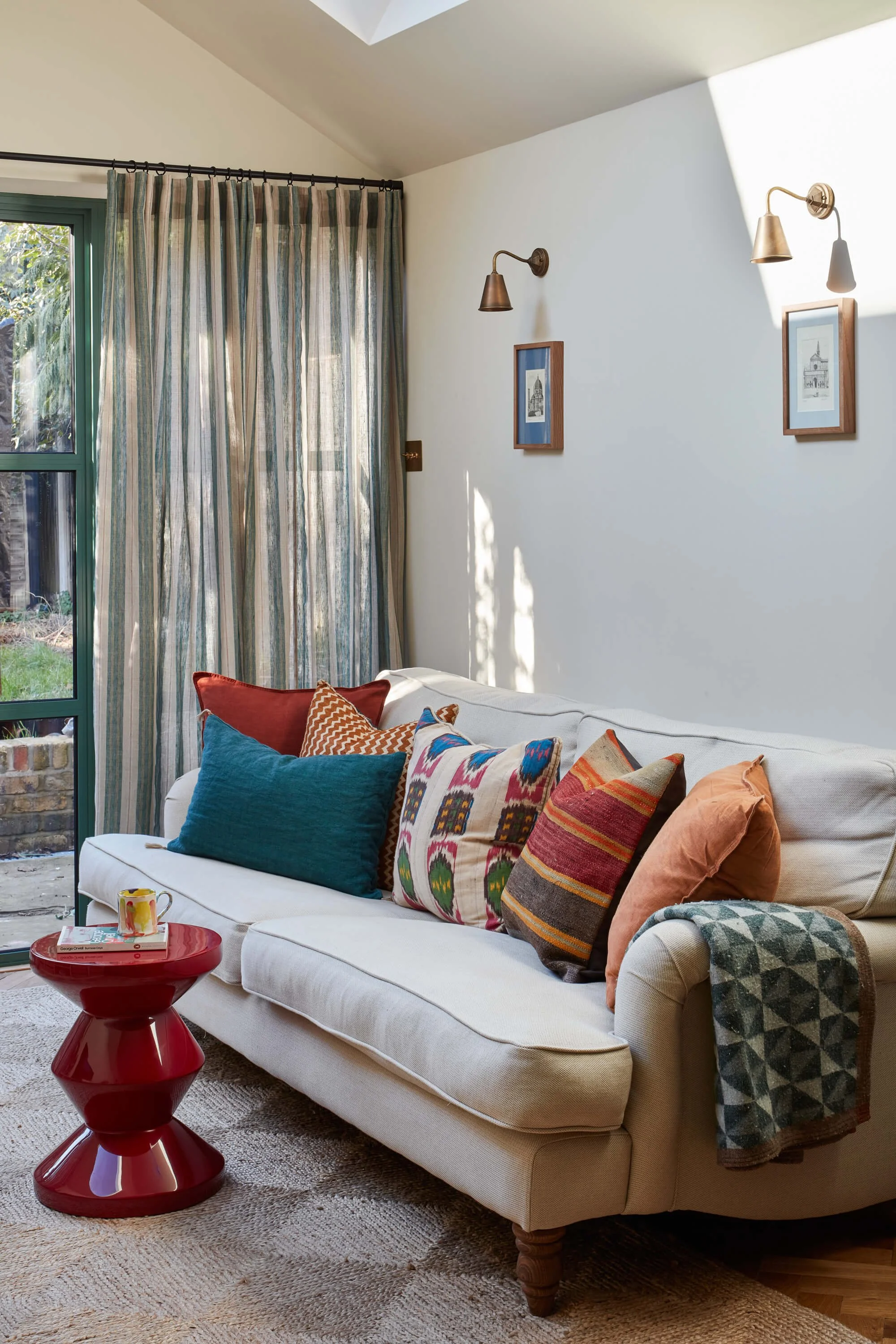 A cozy living room with a white couch decorated with colorful pillows, a red side table with books and a mug, a window with green-framed panes, cream-colored walls with framed art, brass wall-mounted lamps, striped curtains, and a textured beige rug.