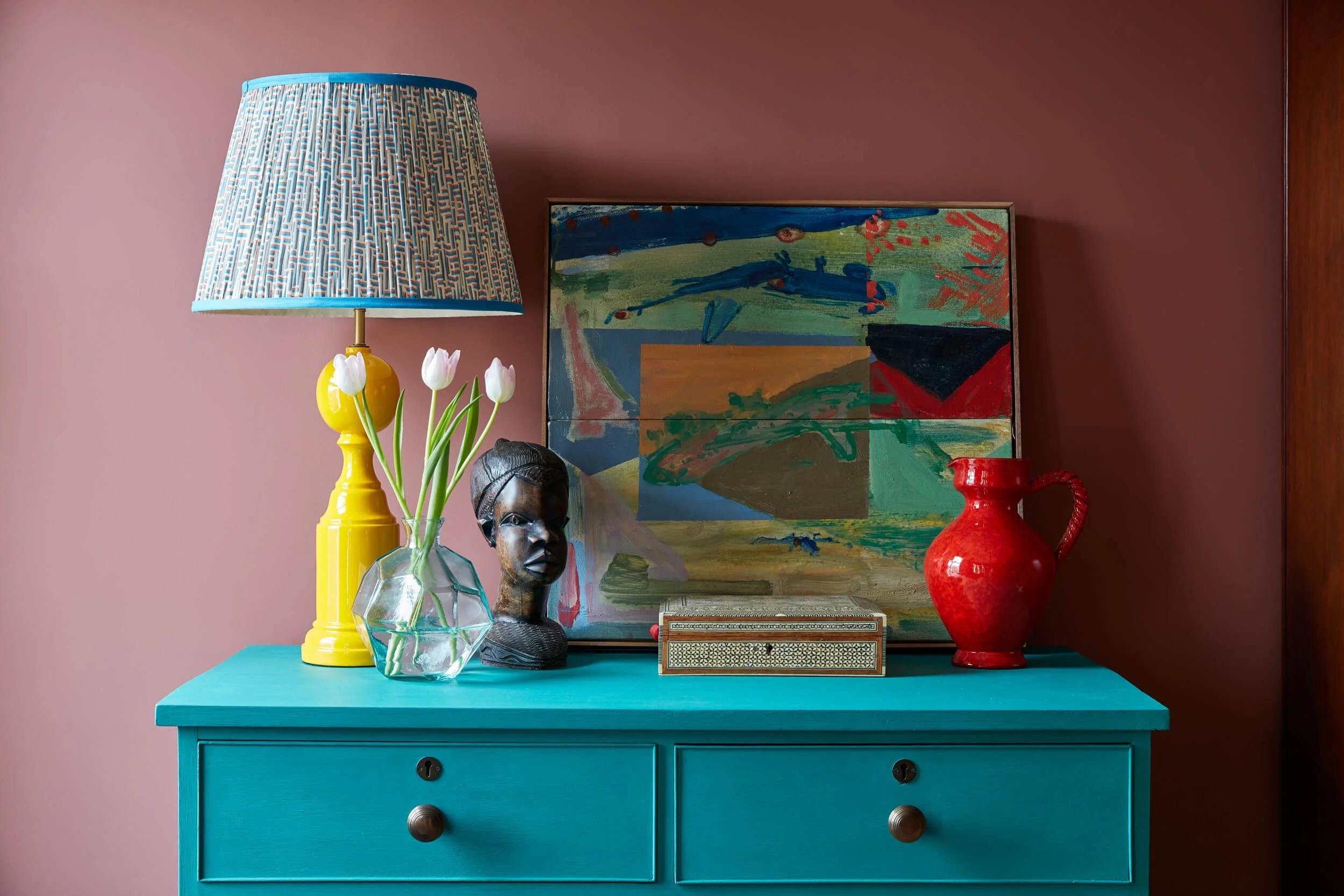 Colorful interior decor featuring a bright turquoise dresser with two drawers, a yellow table lamp with a blue patterned lampshade, a clear faceted glass vase with white tulips, a wooden sculpture, an abstract colorful painting, a small decorative box, a red pitcher, and a red vase, set against a pink wall.