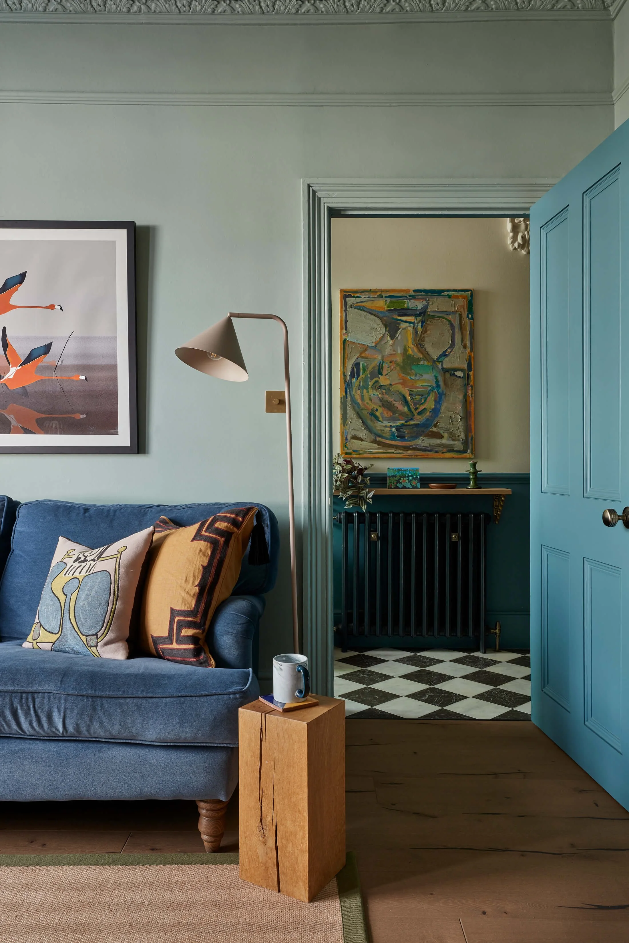 Living room with a blue velvet sofa, decorative pillows, a standing lamp, and a wooden side table with a mug. Part of a painted wall, artwork, and an open door revealing a patterned black-and-white floor and additional artwork inside.