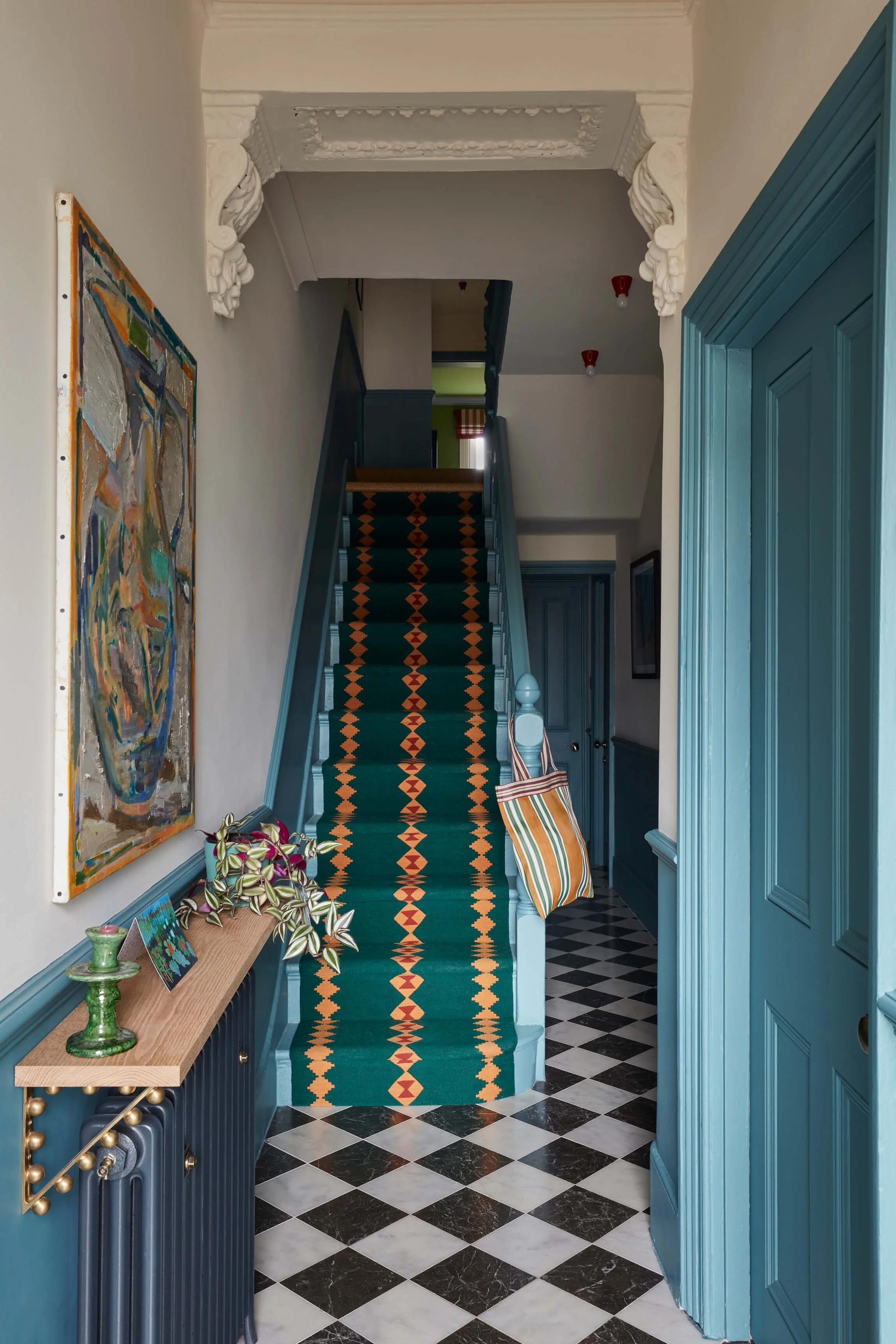 Indoor hallway with black and white checkered floor, blue wainscoting, a staircase with a colorful patterned runner, a small wooden table with plants, paintings, and decorative items, and an open blue door.