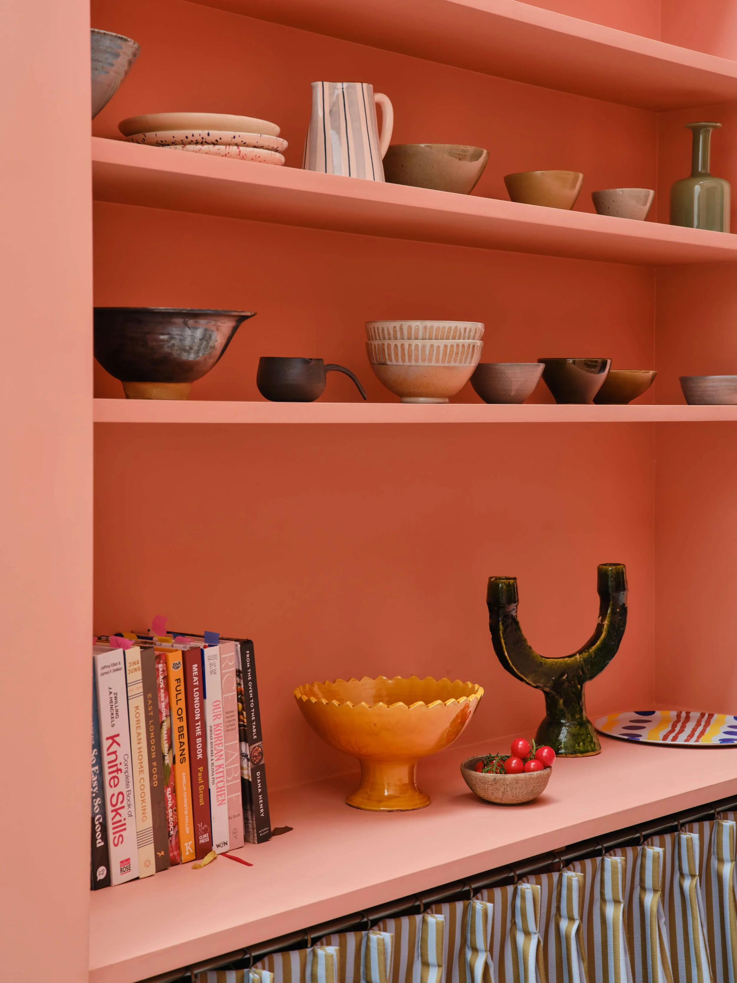 Pink bookshelf with various plates, bowls, a pitcher, a vase, some books, a yellow bowl with a pedestal, and a decorative black candelabra. Curtain with yellow and white stripes hangs at the bottom.