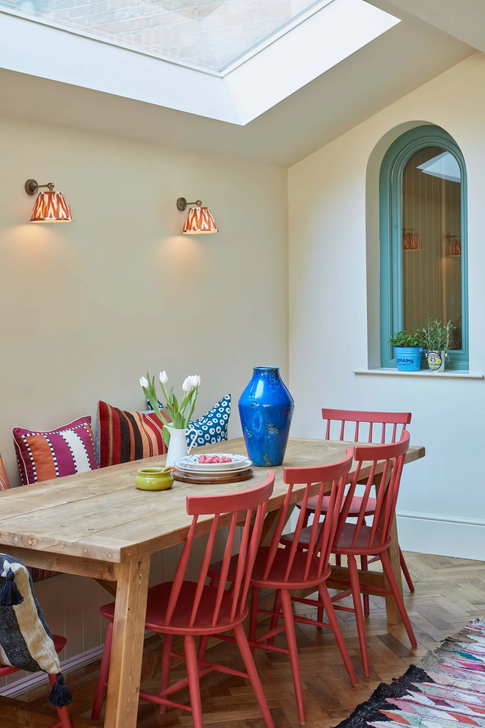 A wooden dining table with pink chairs, colorful pillows on one side, and a vase with white tulips and small bowls on the table. The background has a wall with two wall-mounted lamps, a window with green trim, and some potted herbs on the windowsill under a skylight.