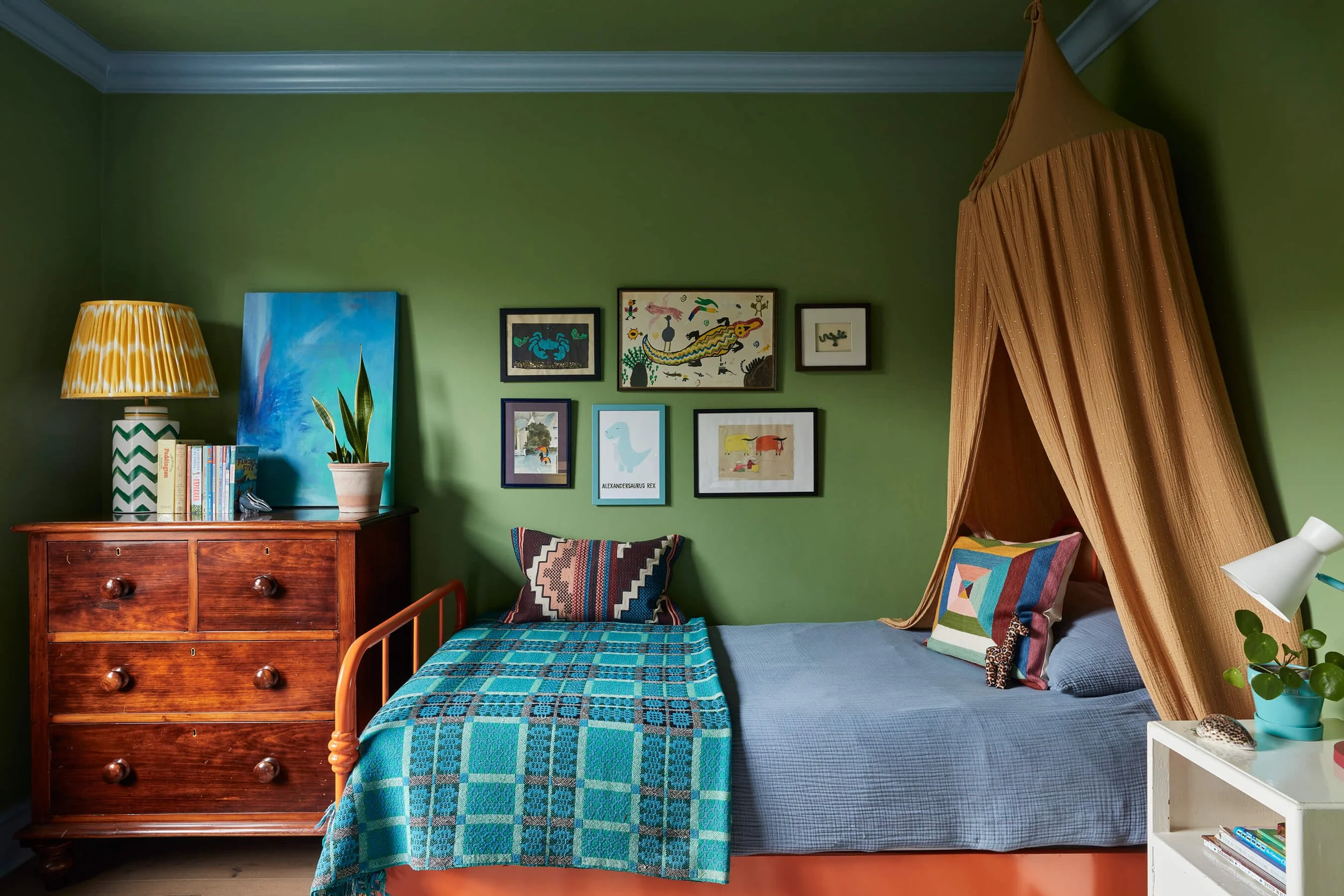 A bedroom with green walls and a wooden dresser, books, a potted plant, a blue painting, and framed pictures on the wall. There is a bed with blue linens, colorful pillows, and a canopy made of orange striped fabric.