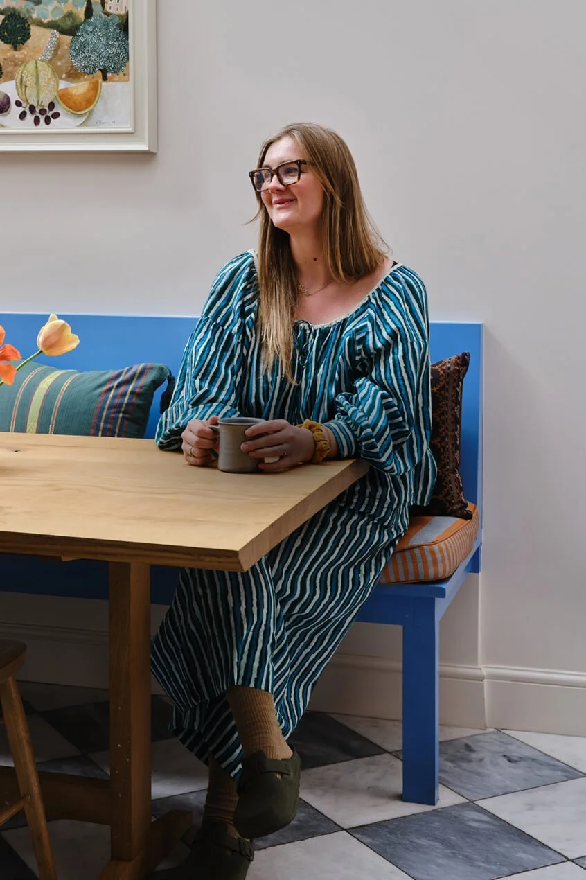A woman with long hair, glasses, and a blue and white striped dress sitting at a wooden table, holding a mug, smiling, in a room with colorful decor and artwork on the wall.