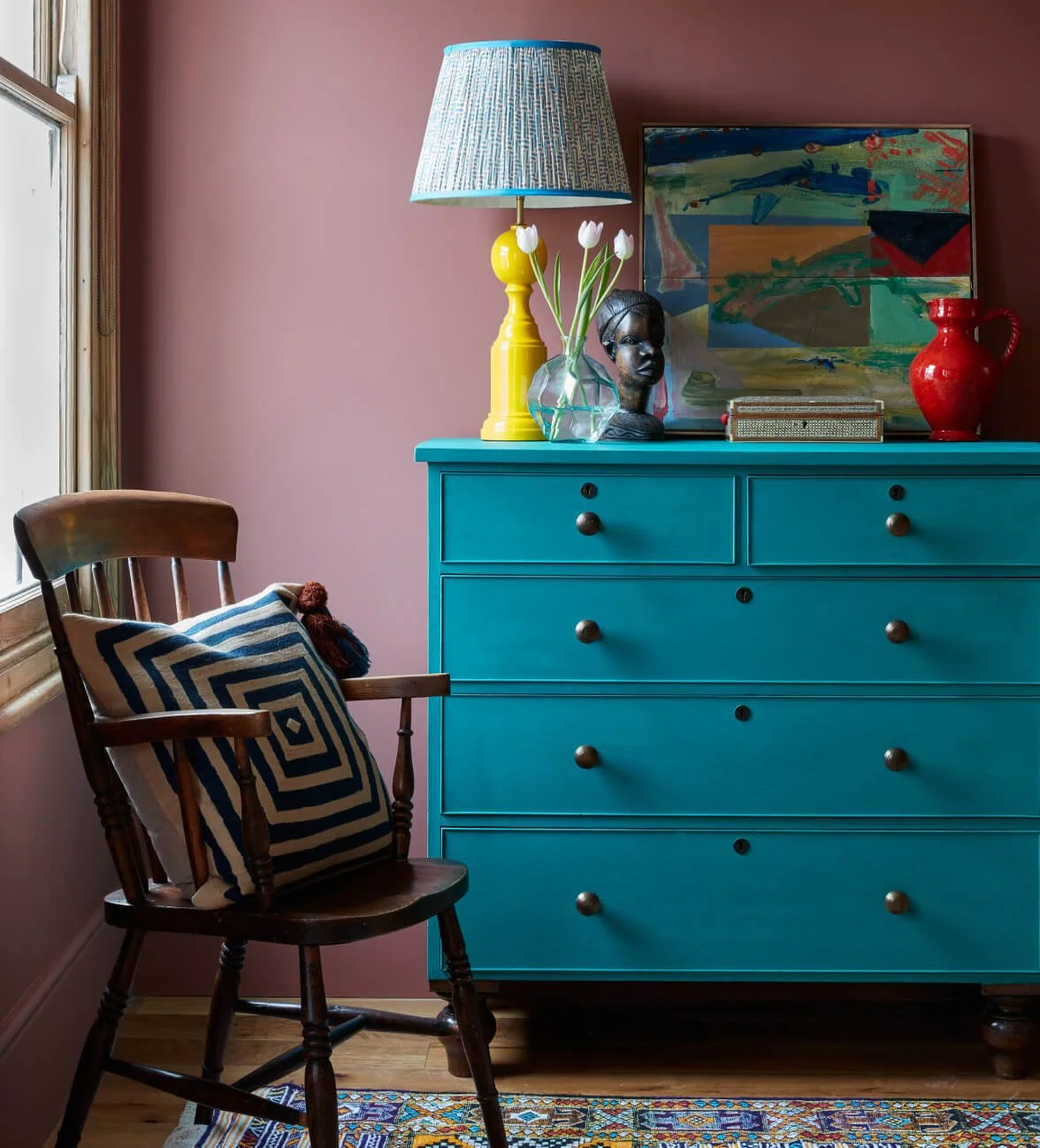 A wooden armchair with a patterned cushion is next to a blue dresser decorated with a yellow lamp, white tulips in a glass vase, a black bust sculpture, a colorful abstract painting, and a red pitcher, against a pink wall and a window.