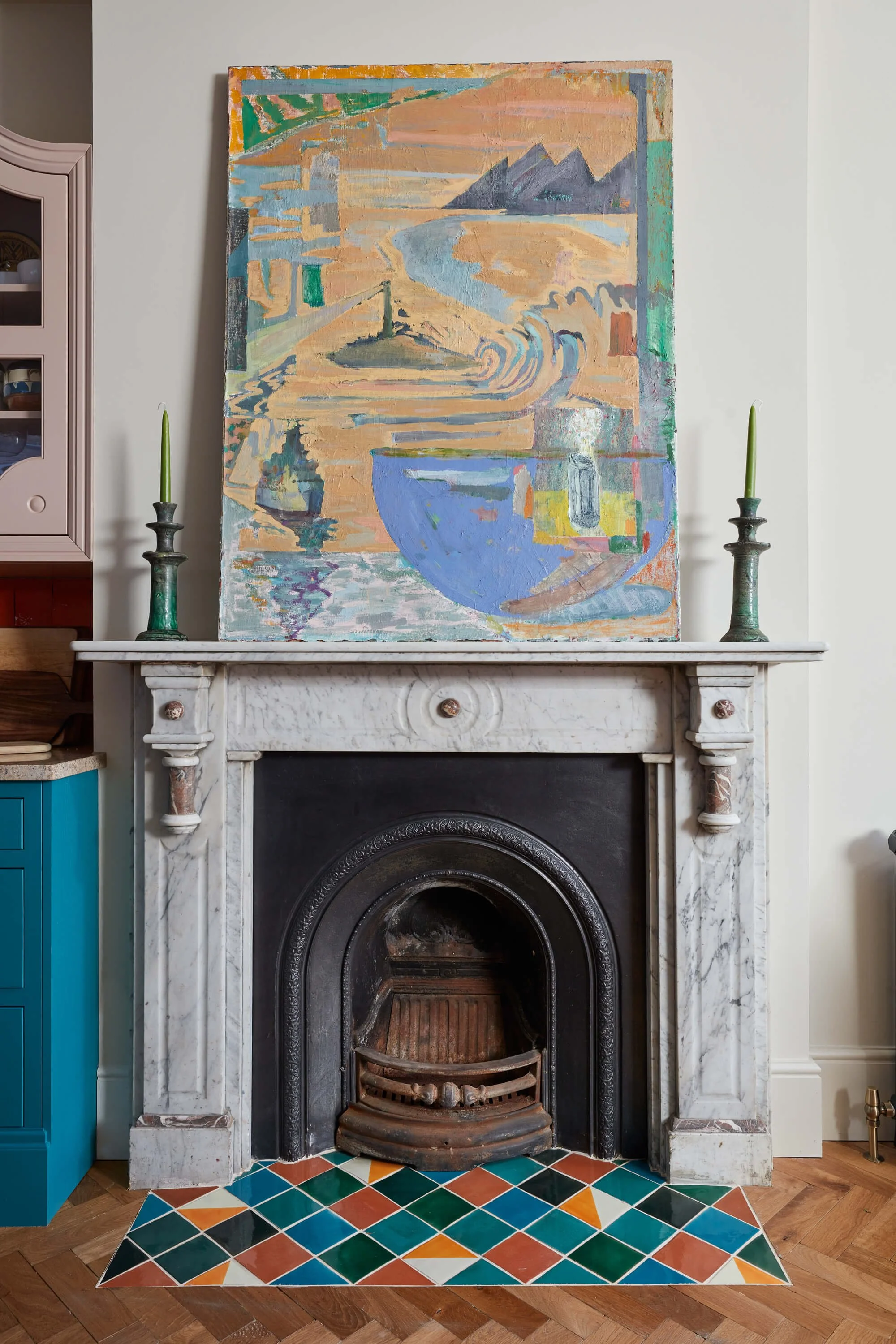 A white fireplace with a marble surround, featuring a painting on the wall above and two green candlesticks on either side. The painting depicts an abstract landscape with mountains, water, and a large blue bowl or vessel. The floor in front of the fireplace has a colorful, geometric tile pattern. Part of a cabinet with dishes is visible to the left.