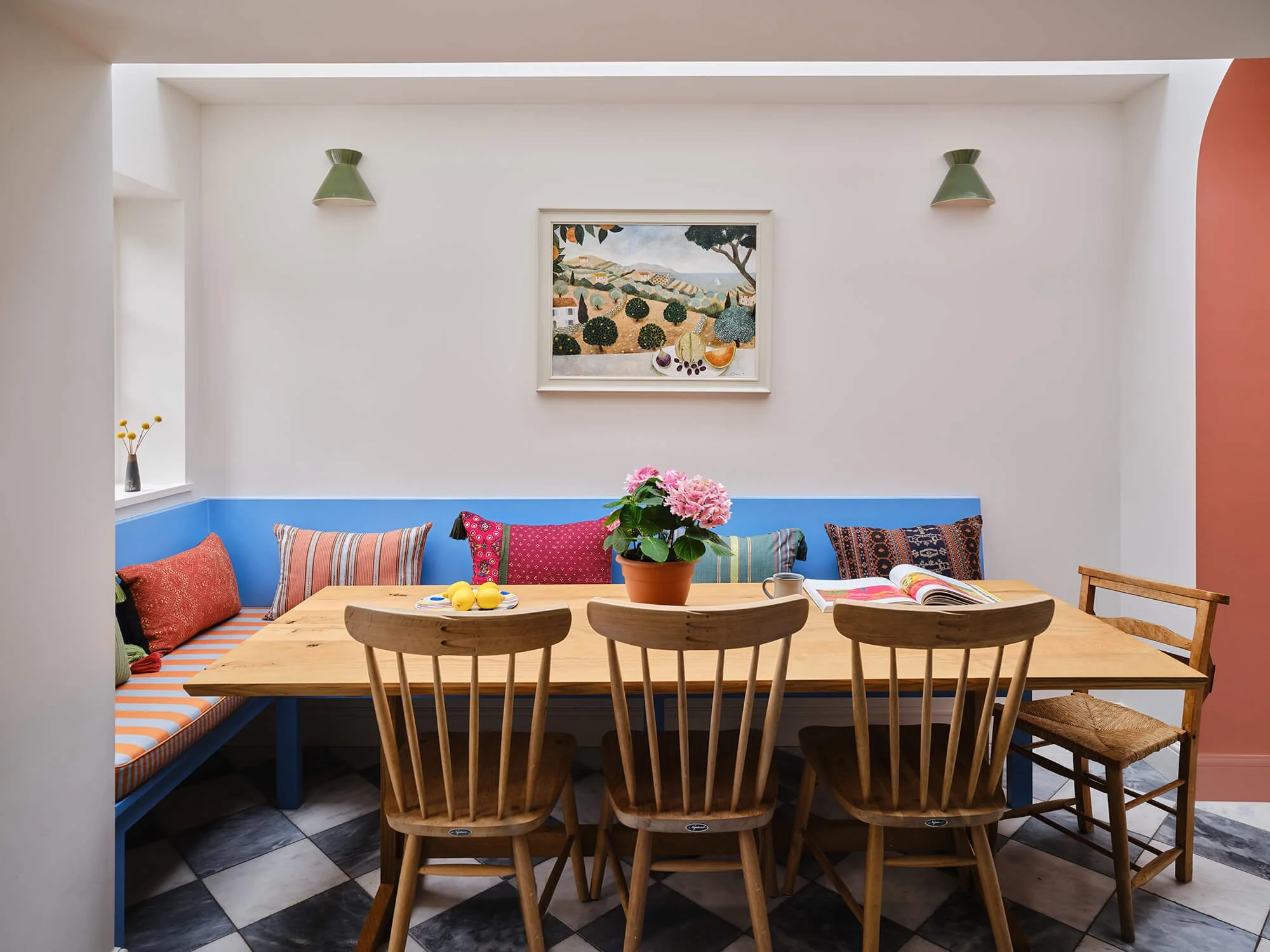 Dining nook with a wooden table, surrounded by wooden chairs and a bench with colorful cushions, with a flower pot and lemons on the table, framed painting on the wall, and green wall sconces.