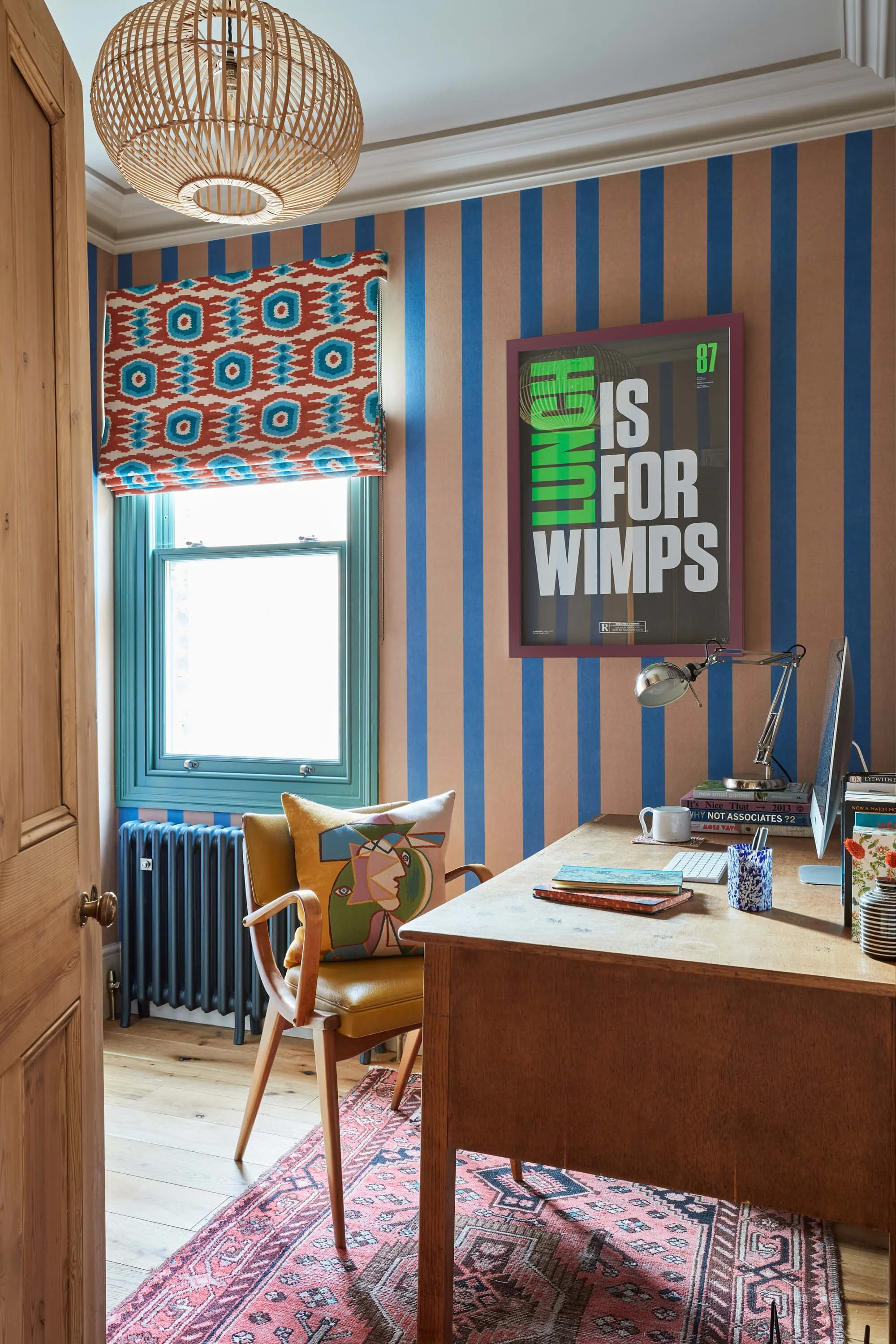 A cozy home office with a wooden desk, a yellow armchair with a colorful pillow, a window with a patterned Roman shade, a framed poster reading 'LUNCH IS FOR WIMPS' on the striped wallpaper, a desk lamp, and a stack of books.