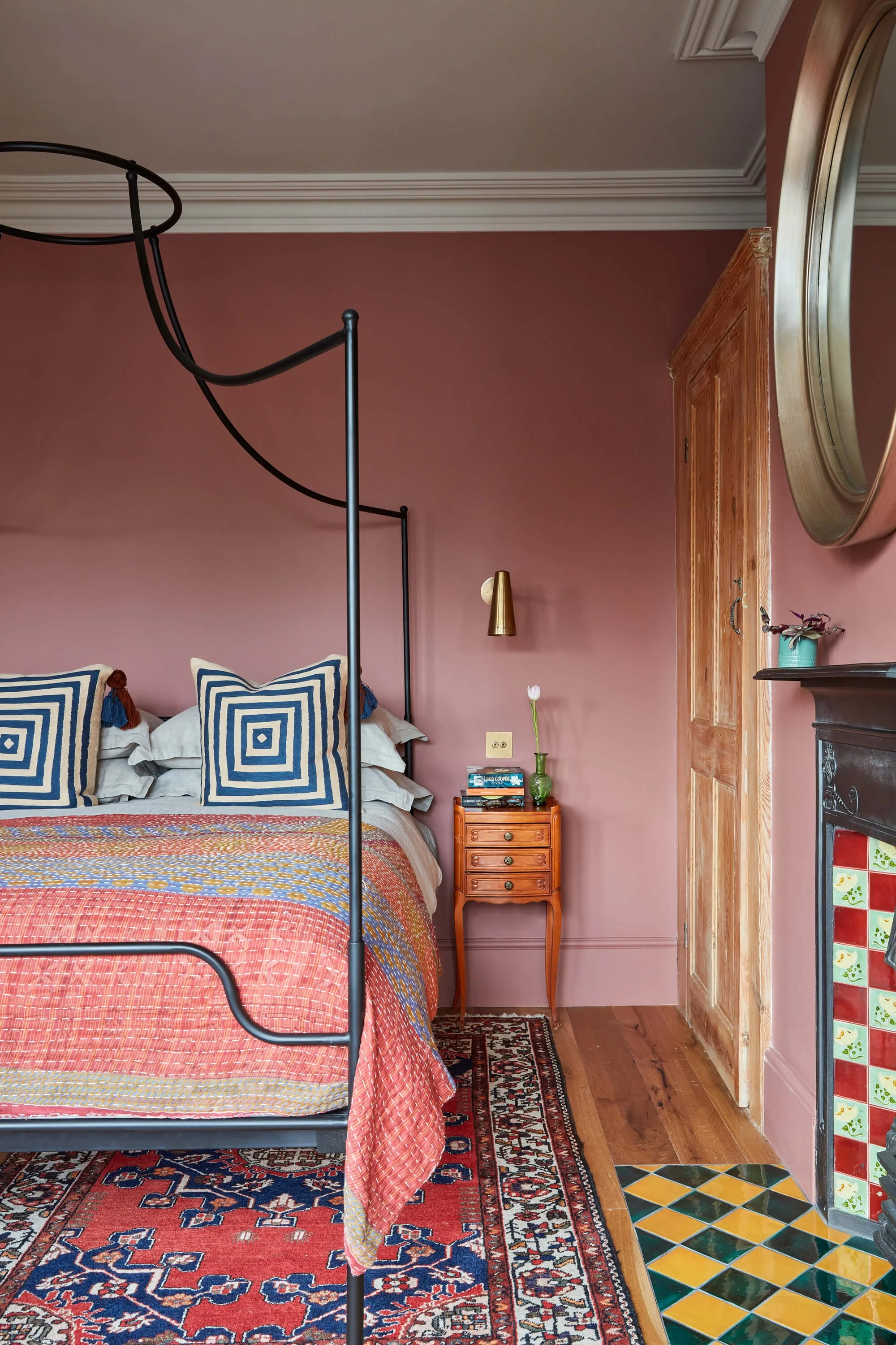 A cozy bedroom with a four-poster bed featuring blue and white geometric pillows and a colorful quilt. Beside the bed is a small wooden nightstand with books and a green vase with a single white tulip. The wall is painted mauve, and there is a gold wall sconce. On the right, a wooden door and a mirror are partially visible. The floor has a colorful patterned rug.
